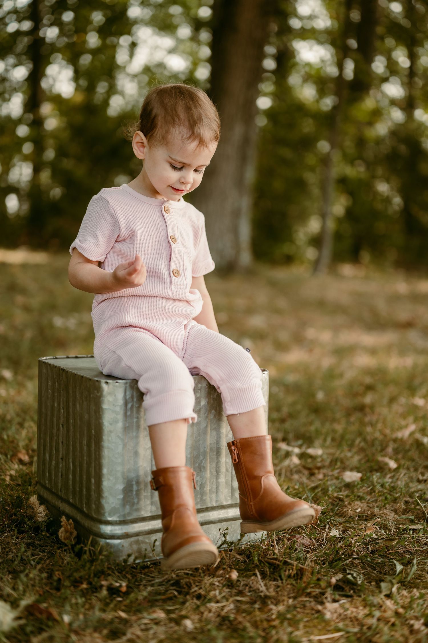 Child in pink outfit sits on vintage metal container in wooded setting.