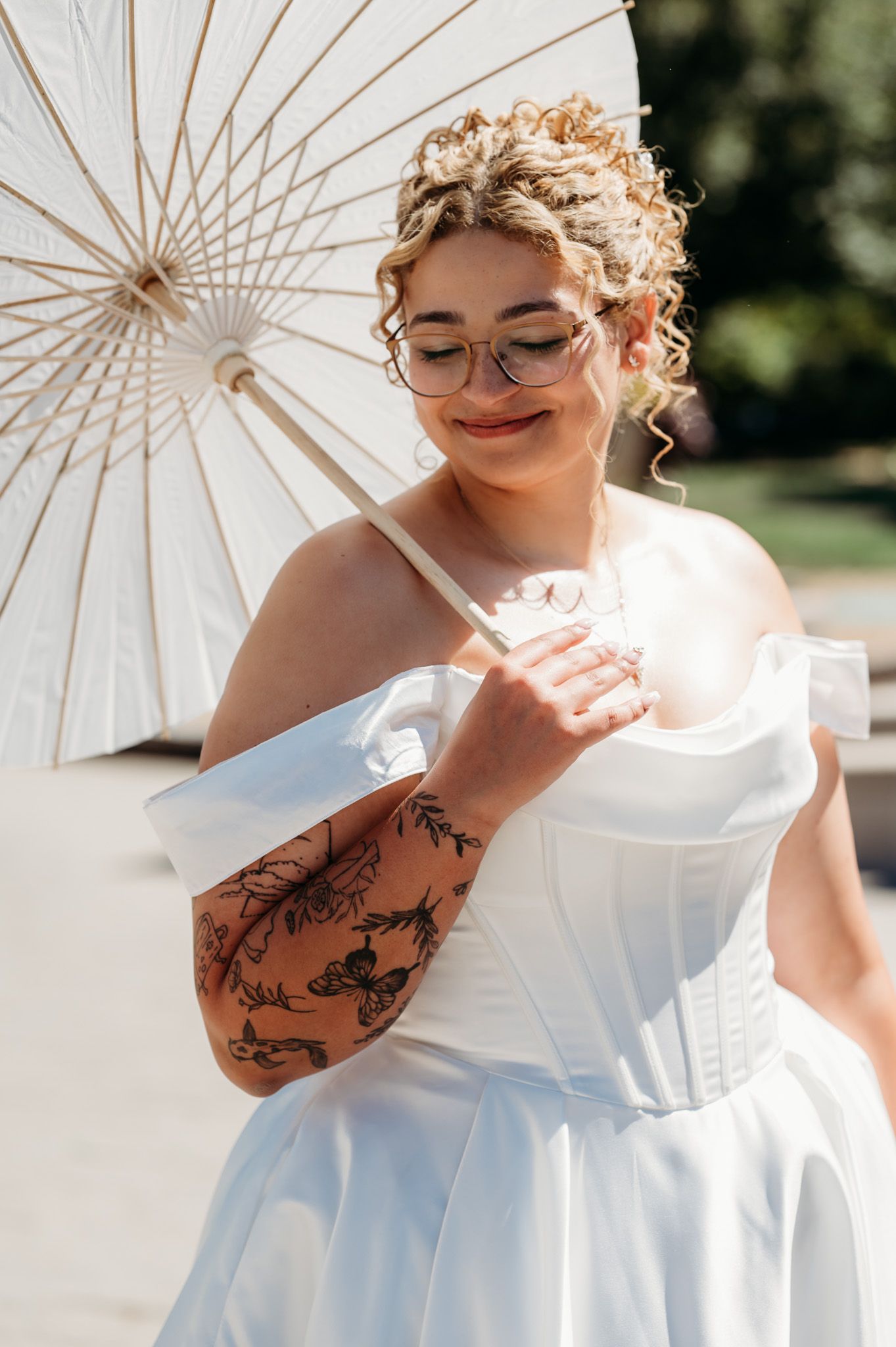 Person in white wedding dress poses with parasol showing artistic tattoo and joyful expression.