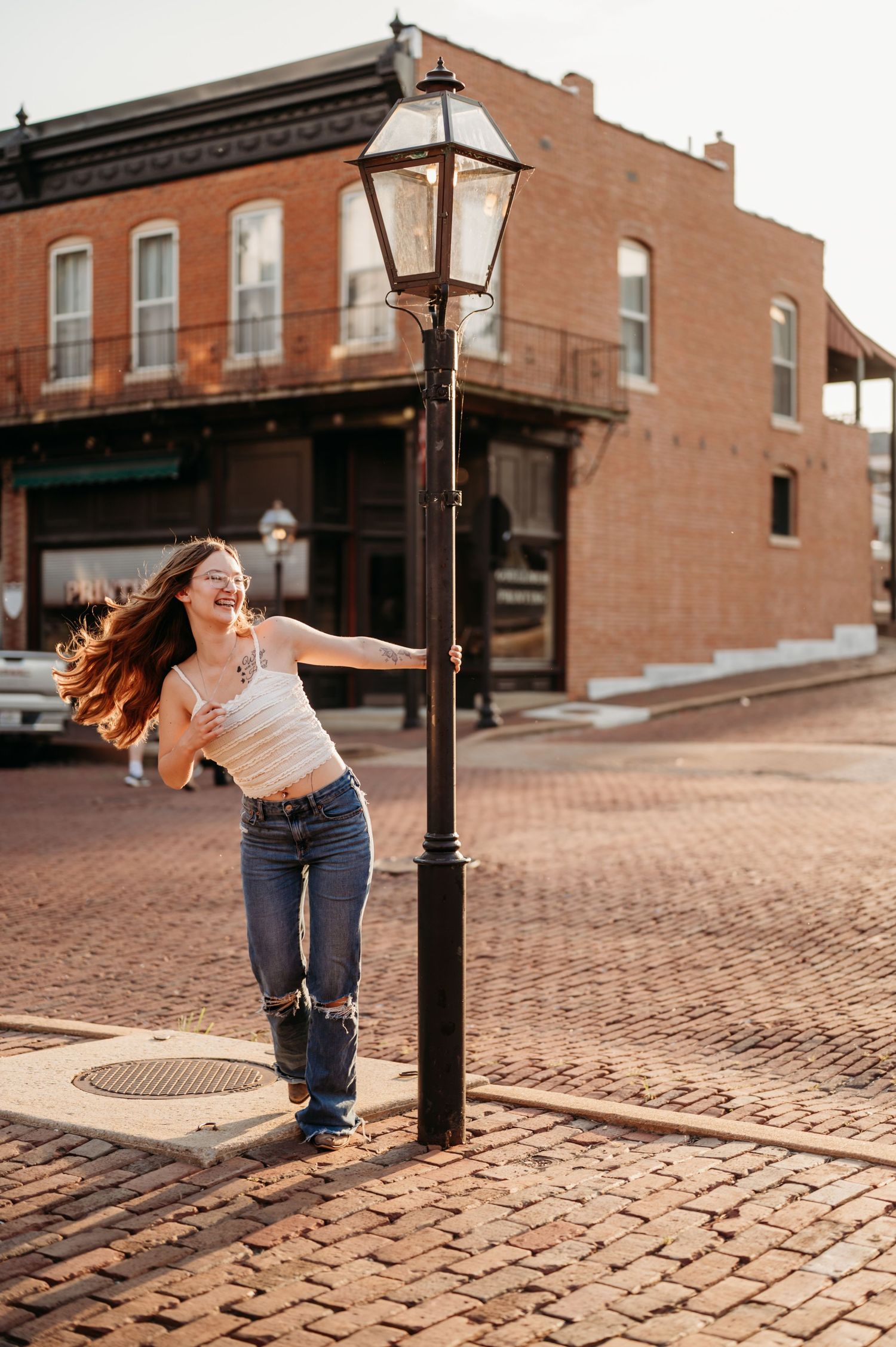 A person in jeans dances and spins around a vintage street lamp post on a brick cobblestone street.