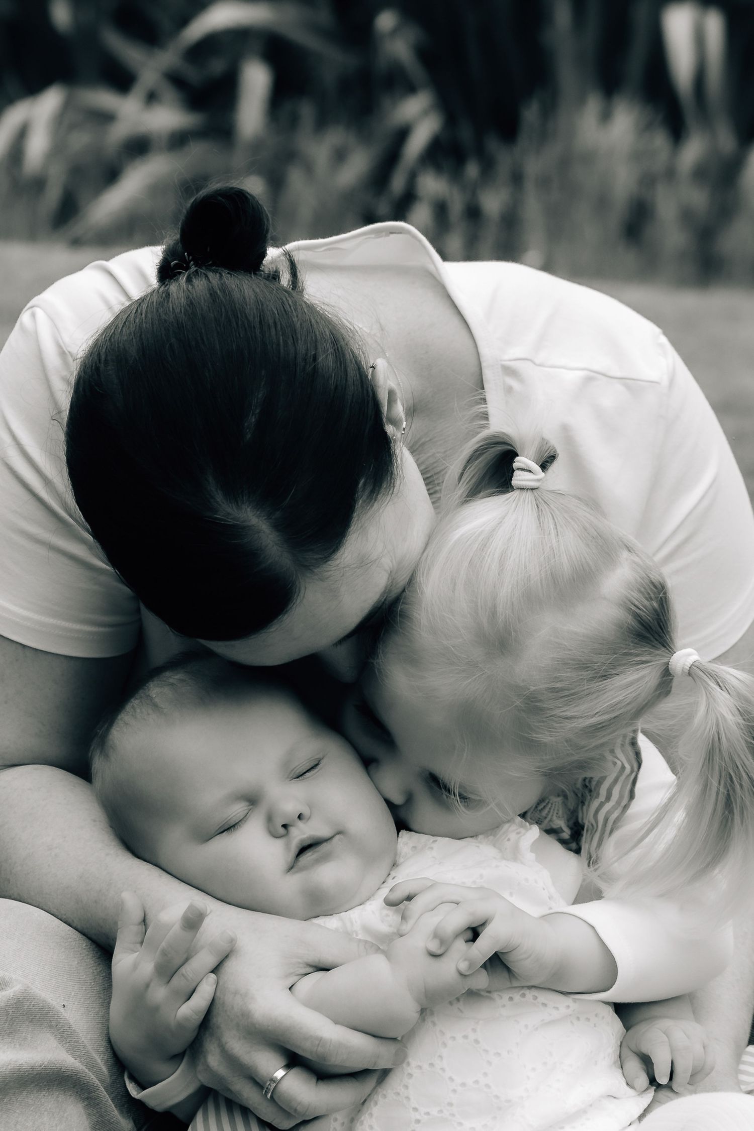 Black and white photo of a mother and toddler kissing a sleeping newborn baby outdoors.