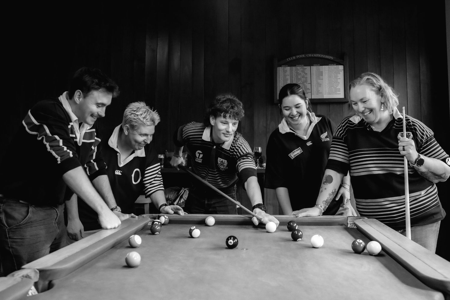 Five people in rugby jerseys laugh while playing pool together in a black and white photo.
