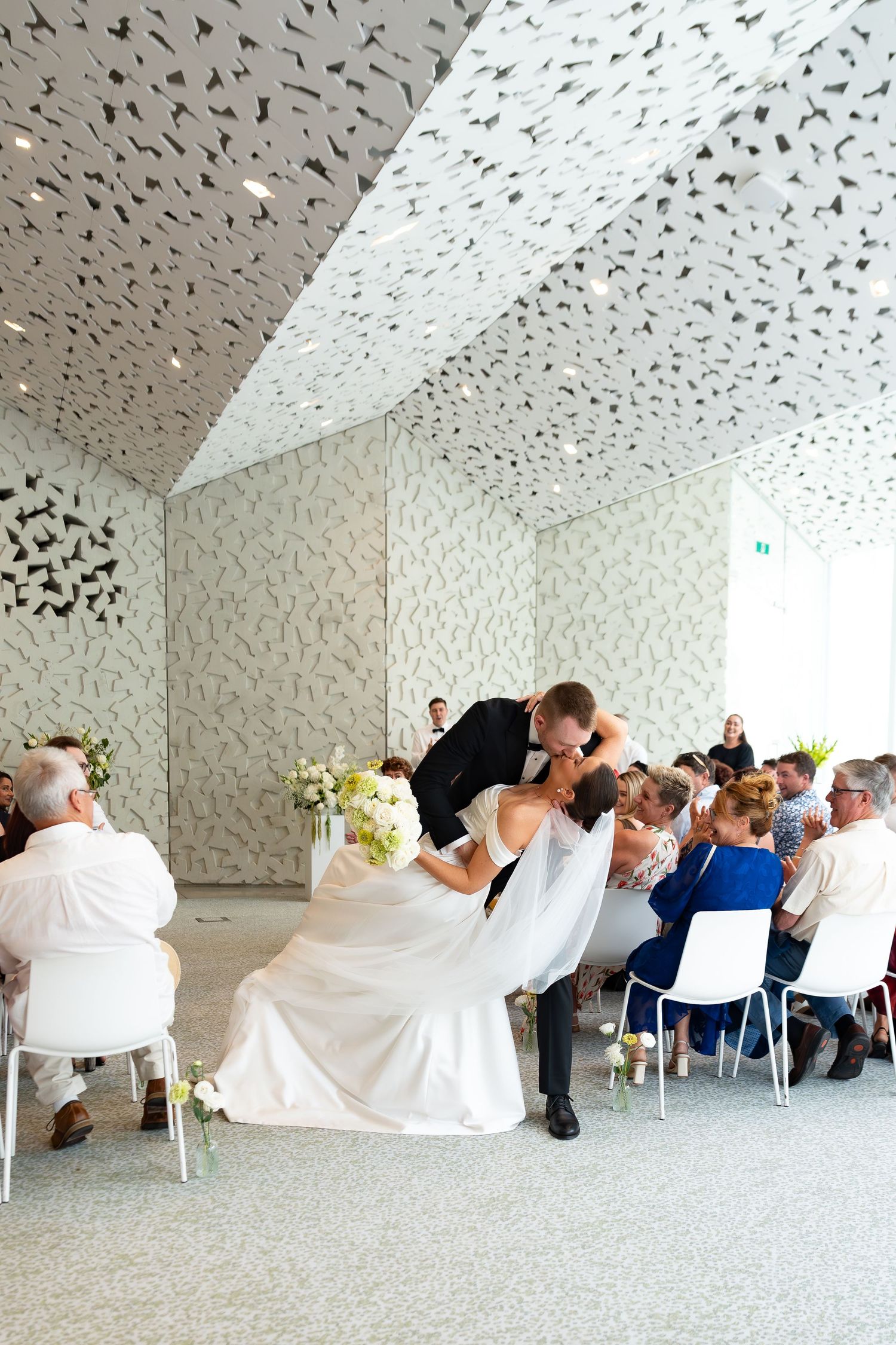 Bride and groom share a romantic dip kiss during their wedding ceremony in a modern white venue with decorative ceiling.