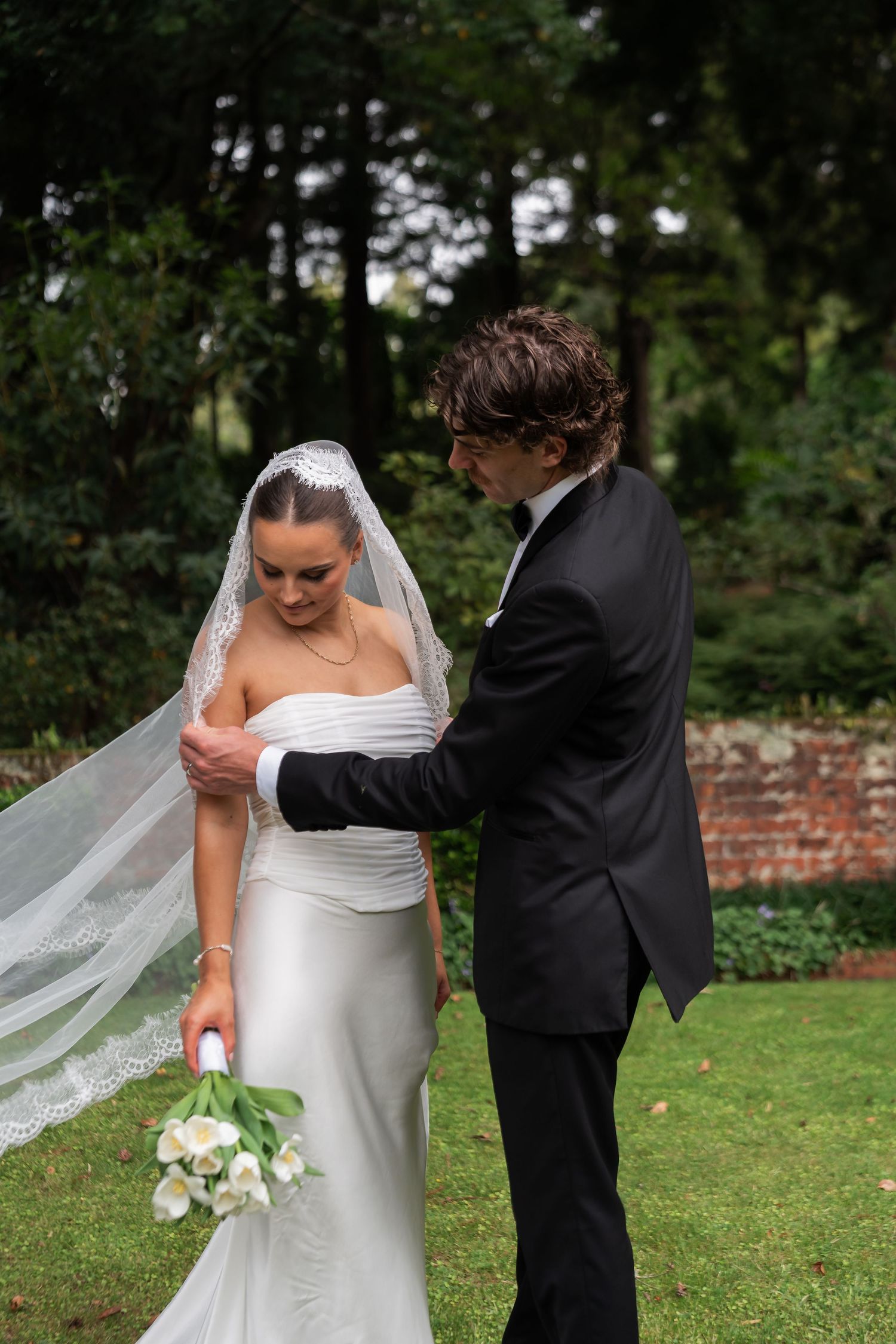 Bride in white gown and lace veil holds tulip bouquet as groom in black tuxedo adjusts her dress outdoors.