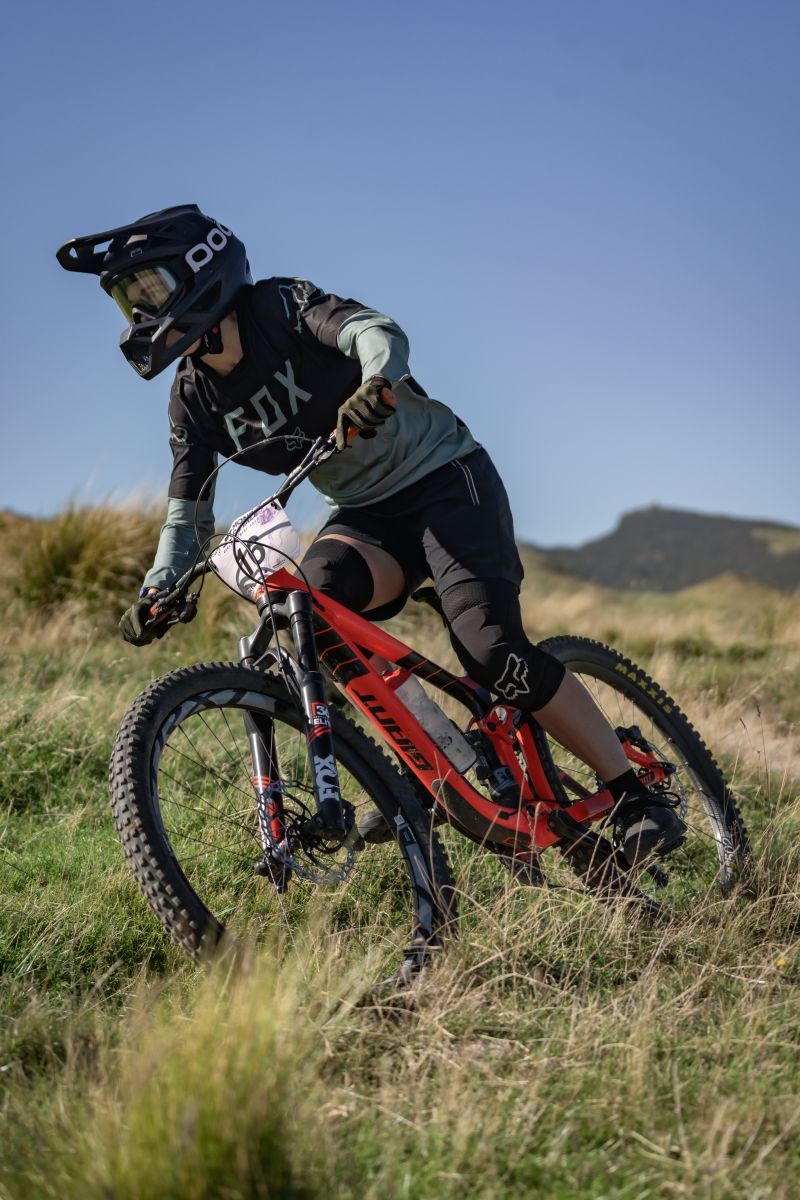 Mountain biker rides red full suspension bike through grassy hillside terrain against blue sky background.