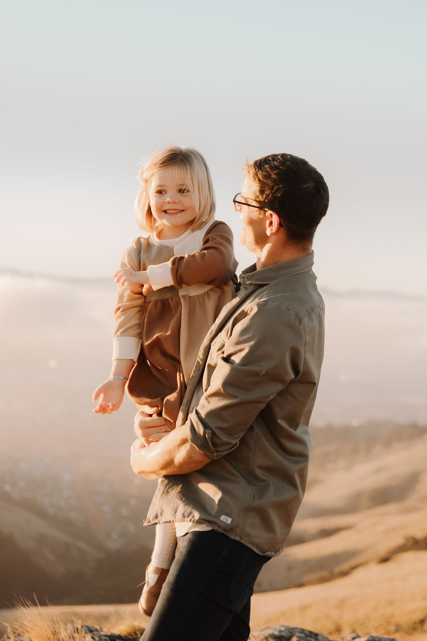 Father holding smiling blonde toddler girl on a sunny hilltop with misty valley views in background.