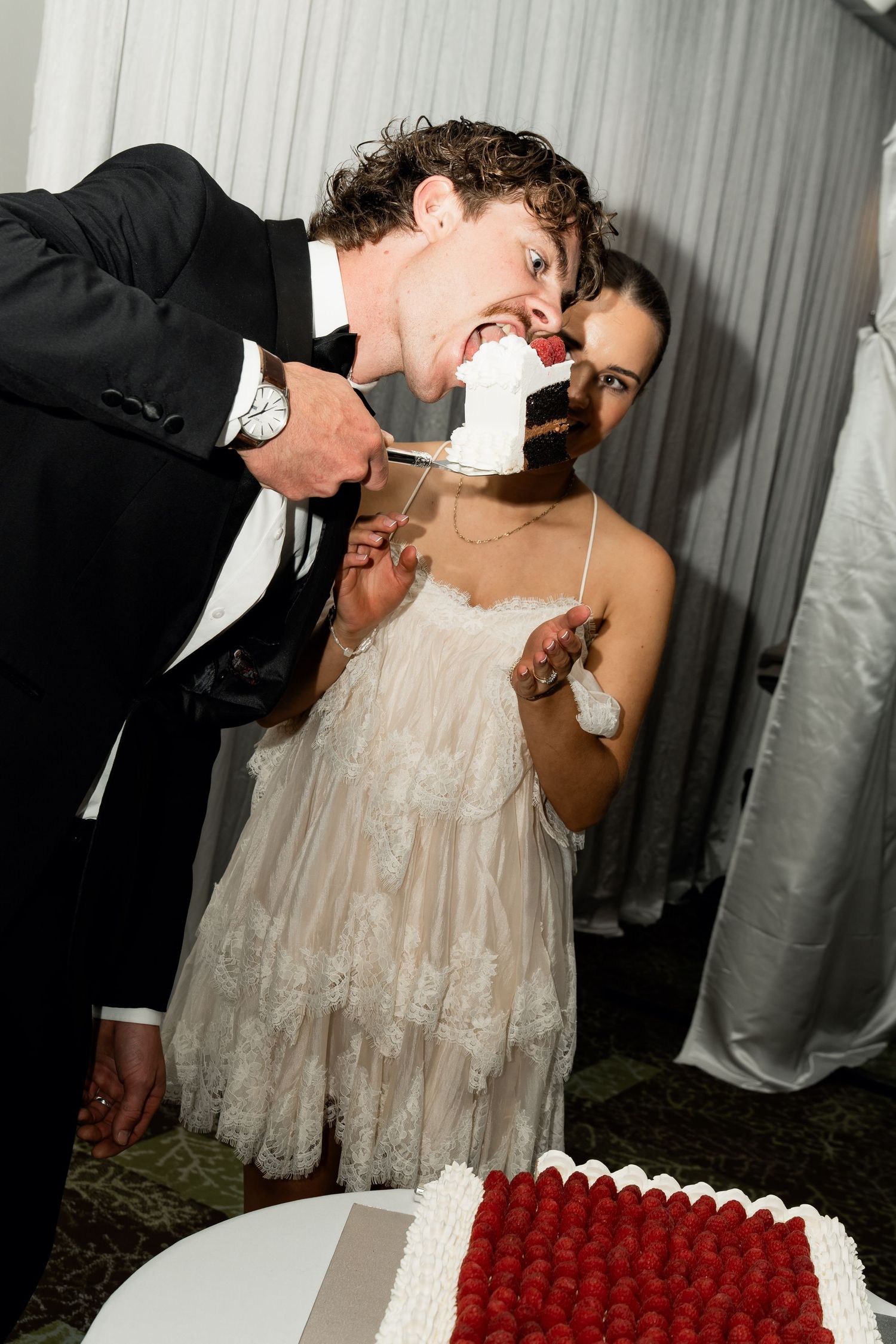 Bride and groom playfully sharing wedding cake, with groom taking a big bite at their elegant reception.