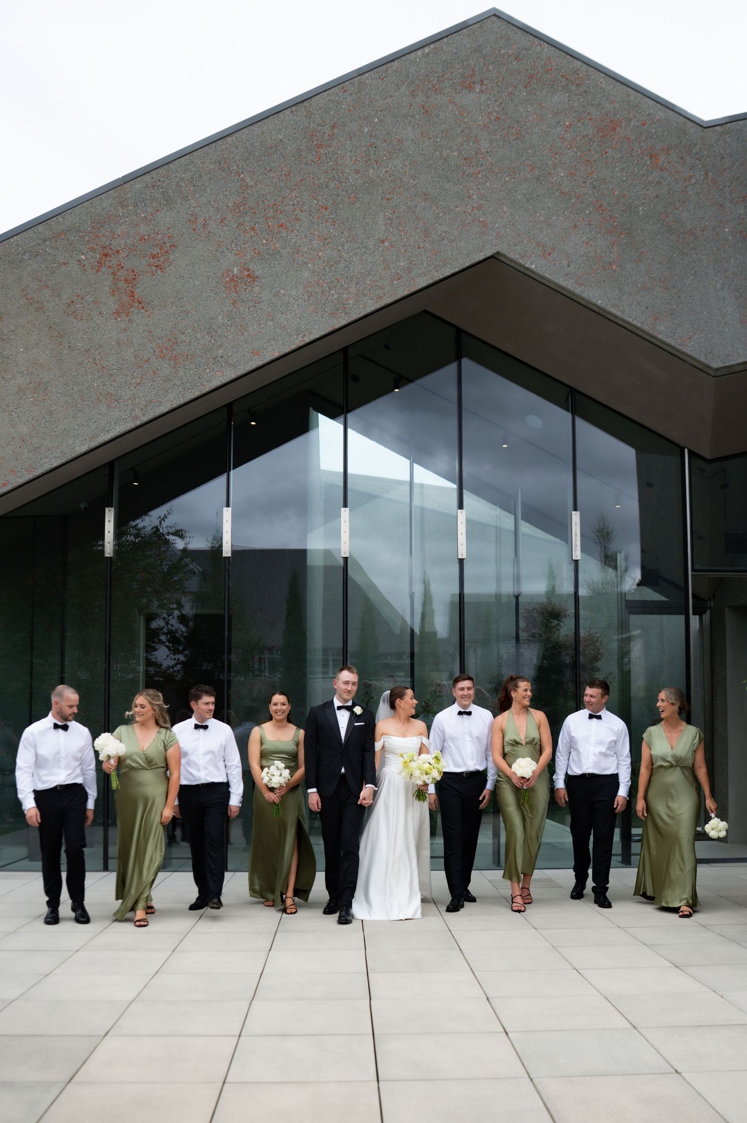 Wedding party poses outside modern glass building, bridesmaids in sage green gowns, groomsmen in black tuxedos.