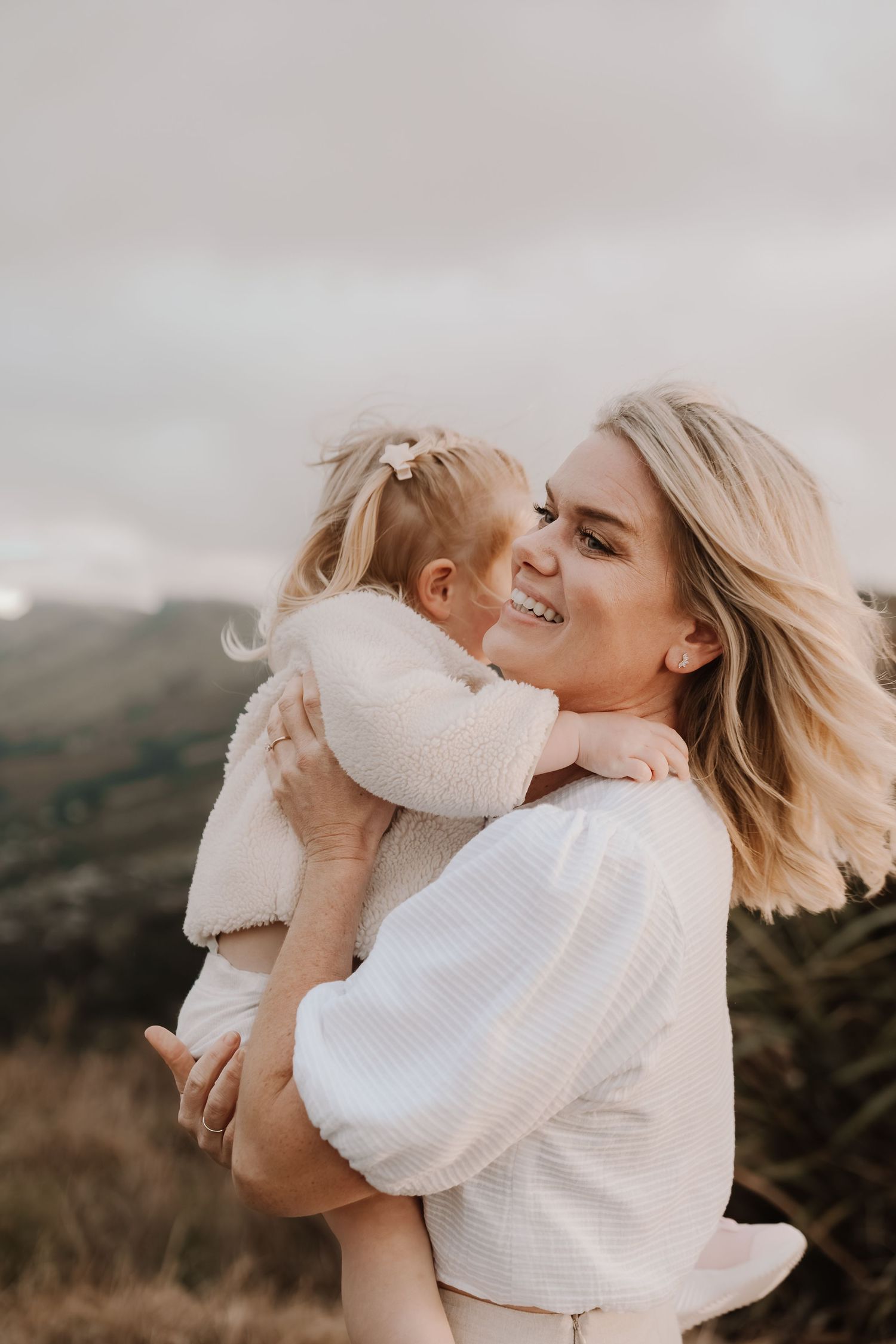 Smiling blonde woman holding a young girl in white outfits, sharing a tender embrace outdoors with hills behind.