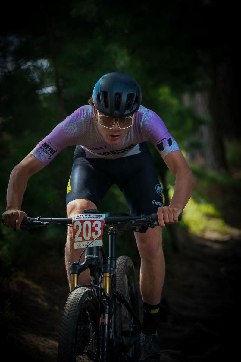 Mountain biker in black cycling gear rides through a wooded trail during a competitive race.