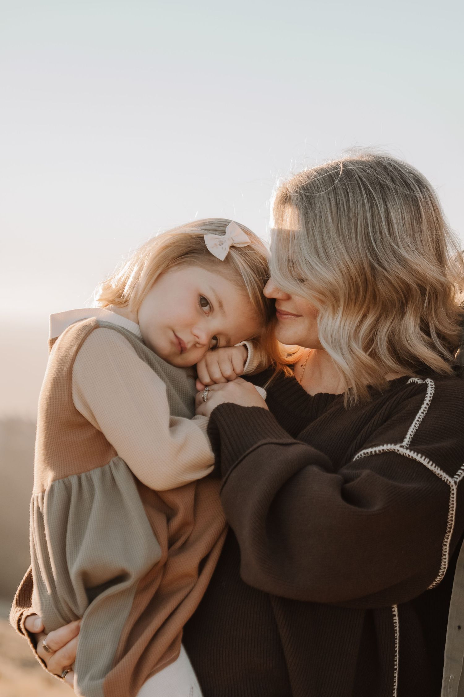 A mother tenderly holds her young daughter close, both sharing a quiet moment outdoors in warm golden light.