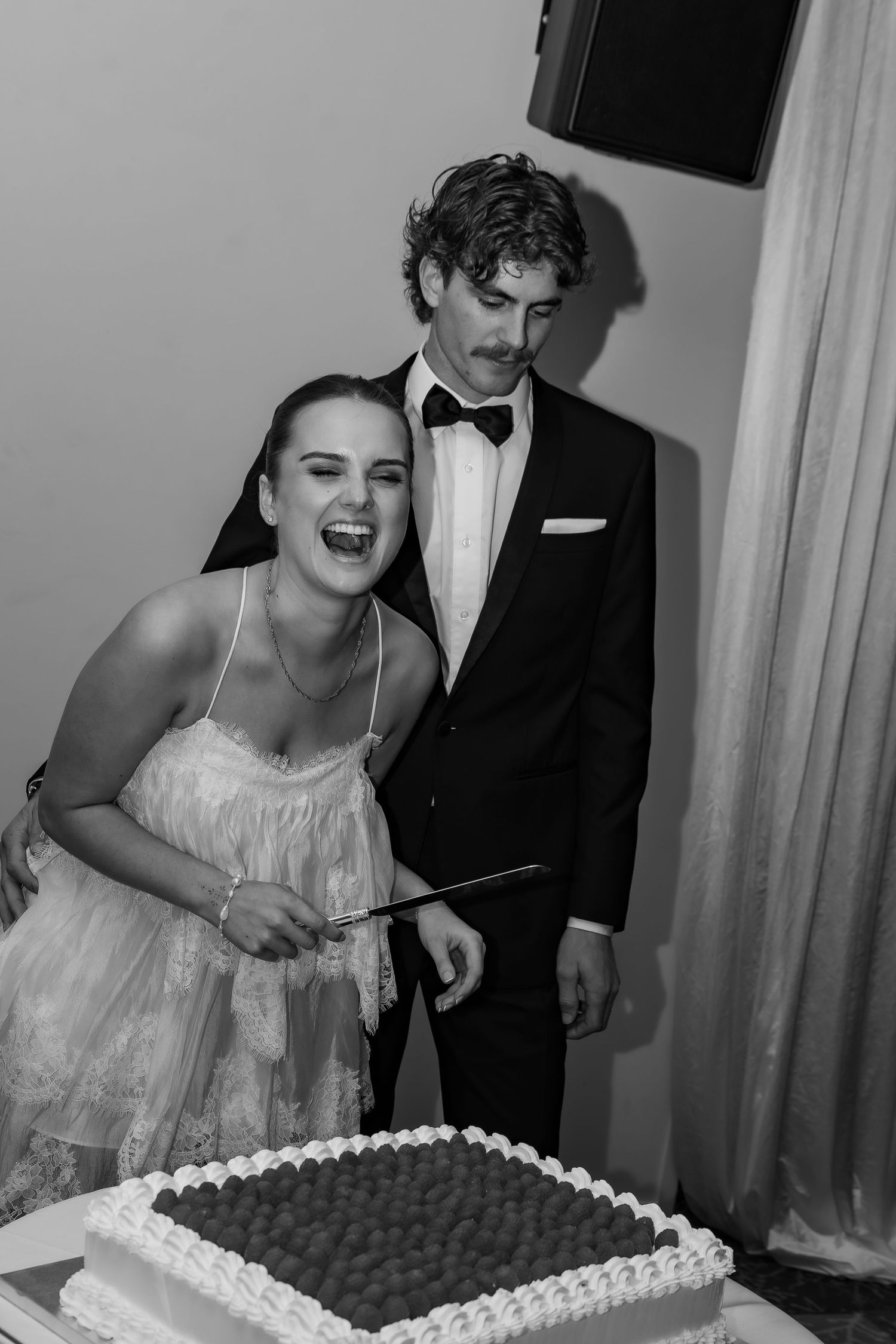 Black and white photo of a bride and groom cutting their wedding cake, bride laughing joyfully.