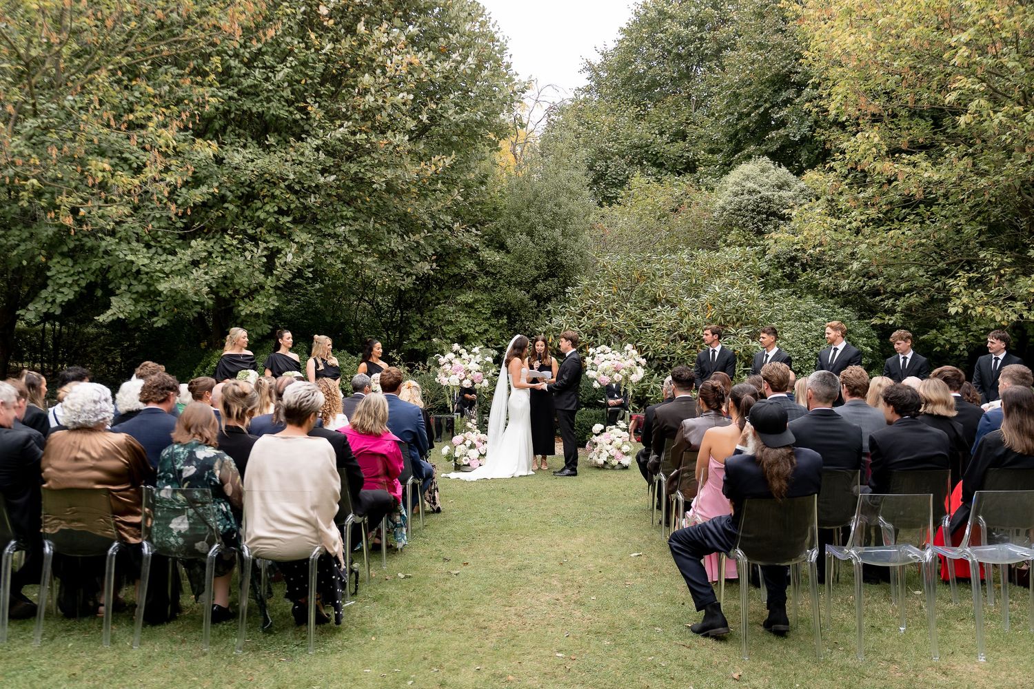 Outdoor wedding ceremony on green lawn with guests seated in rows facing couple at floral arch surrounded by lush trees.