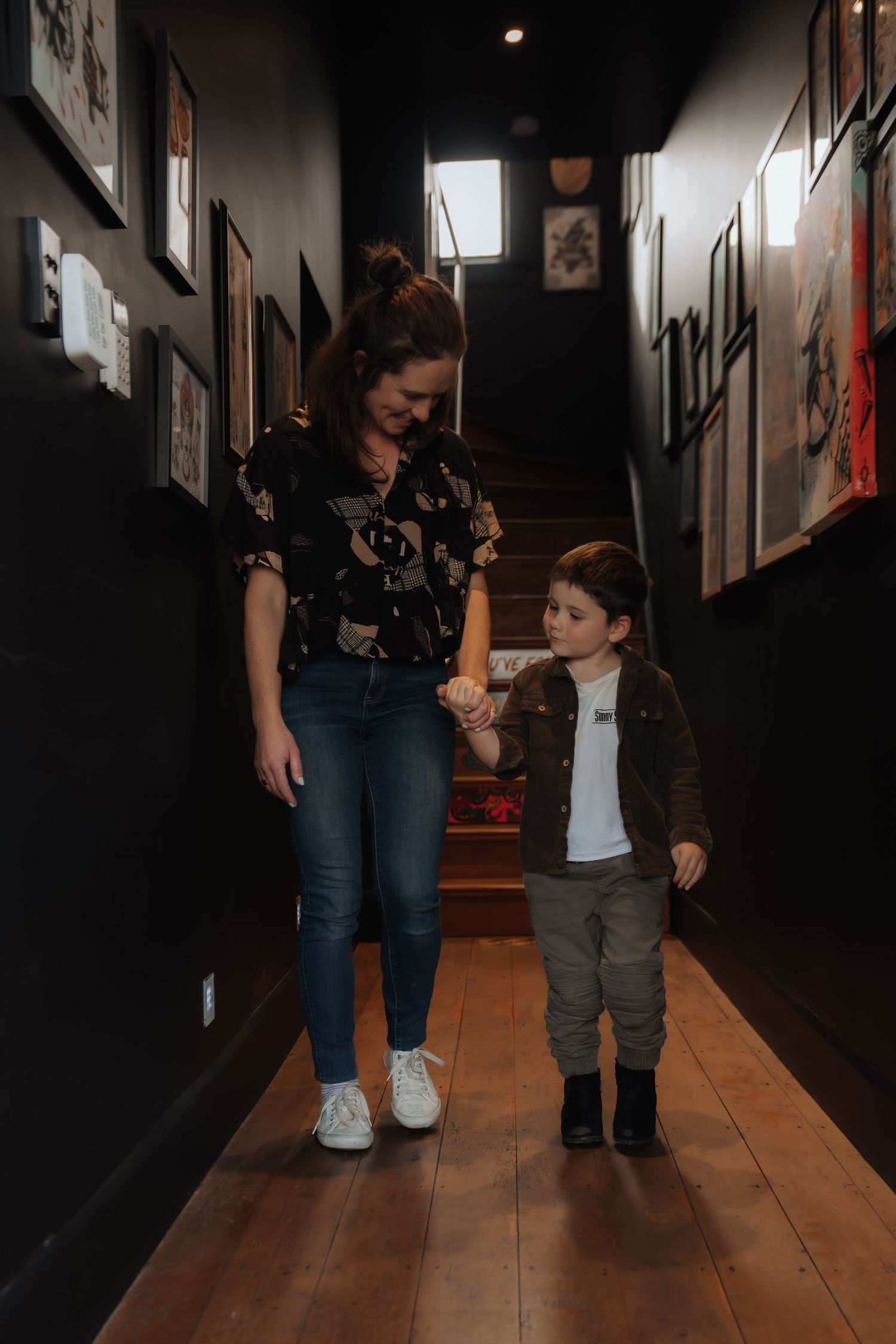 A mother and young child walk hand-in-hand down a dark hallway lined with framed artwork on wooden floors.