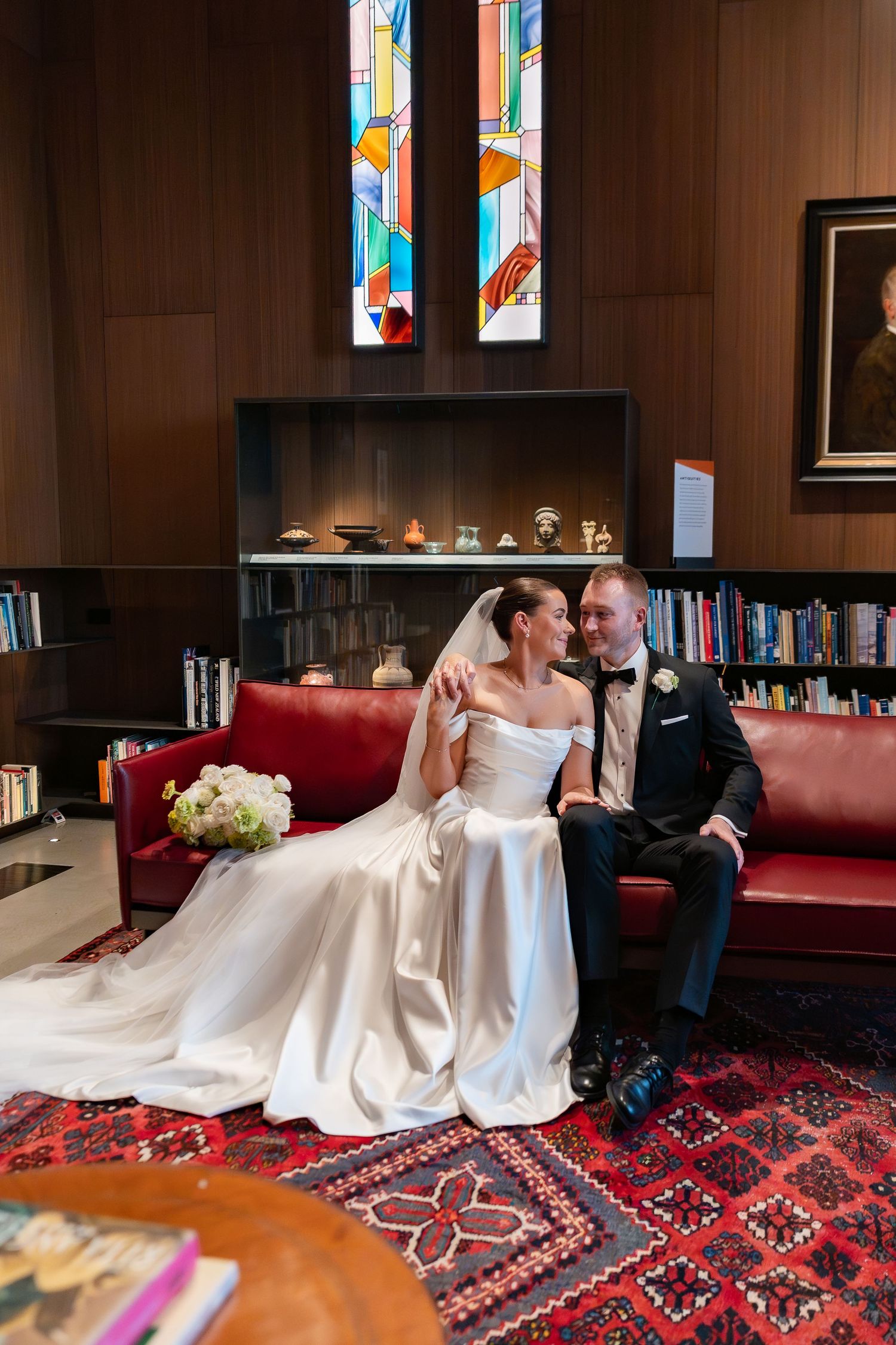 Bride and groom sit together on a red leather sofa in an elegant library with stained glass windows at ravenscar house.