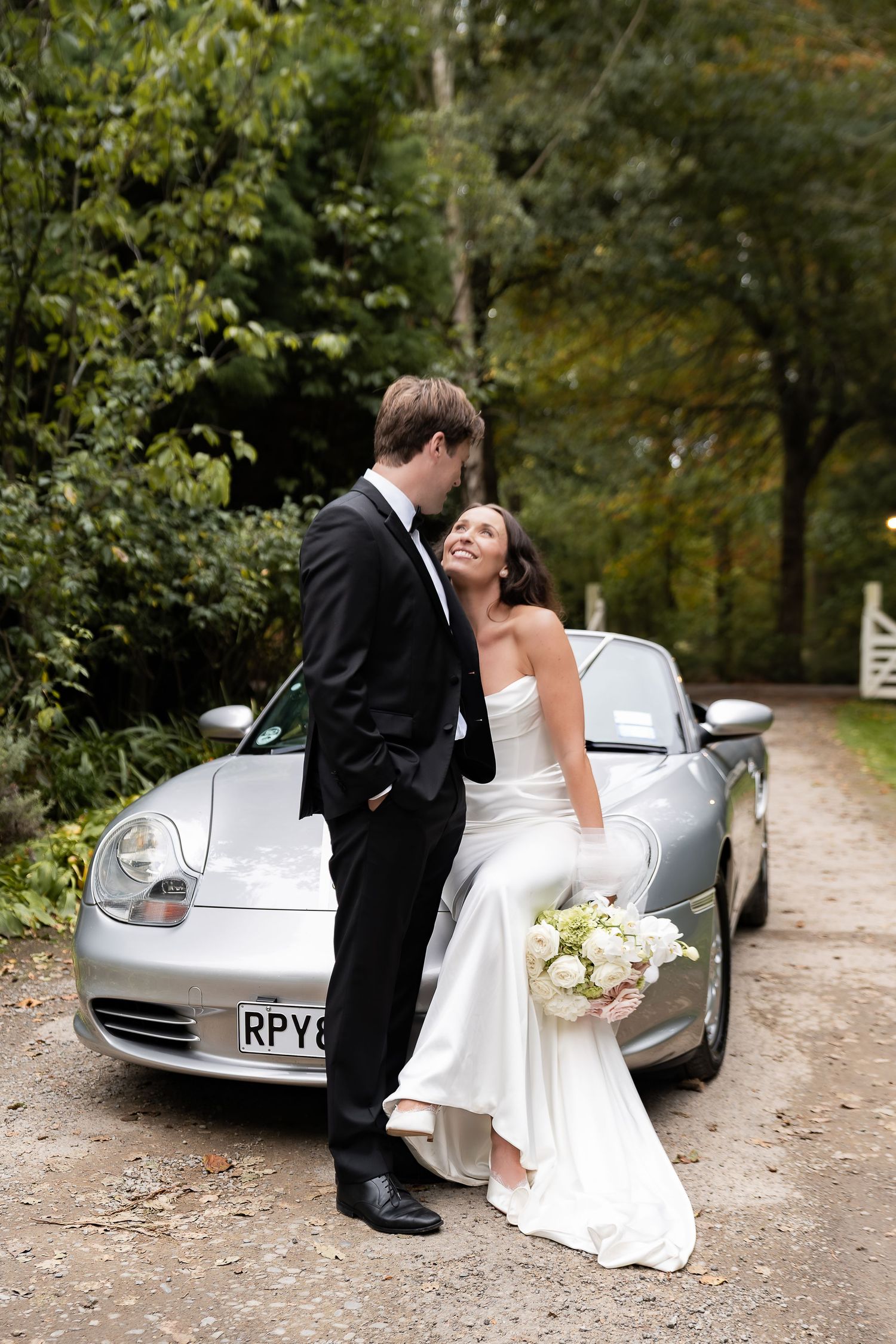A bride and groom pose romantically in front of a silver Porsche on a tree-lined gravel driveway.