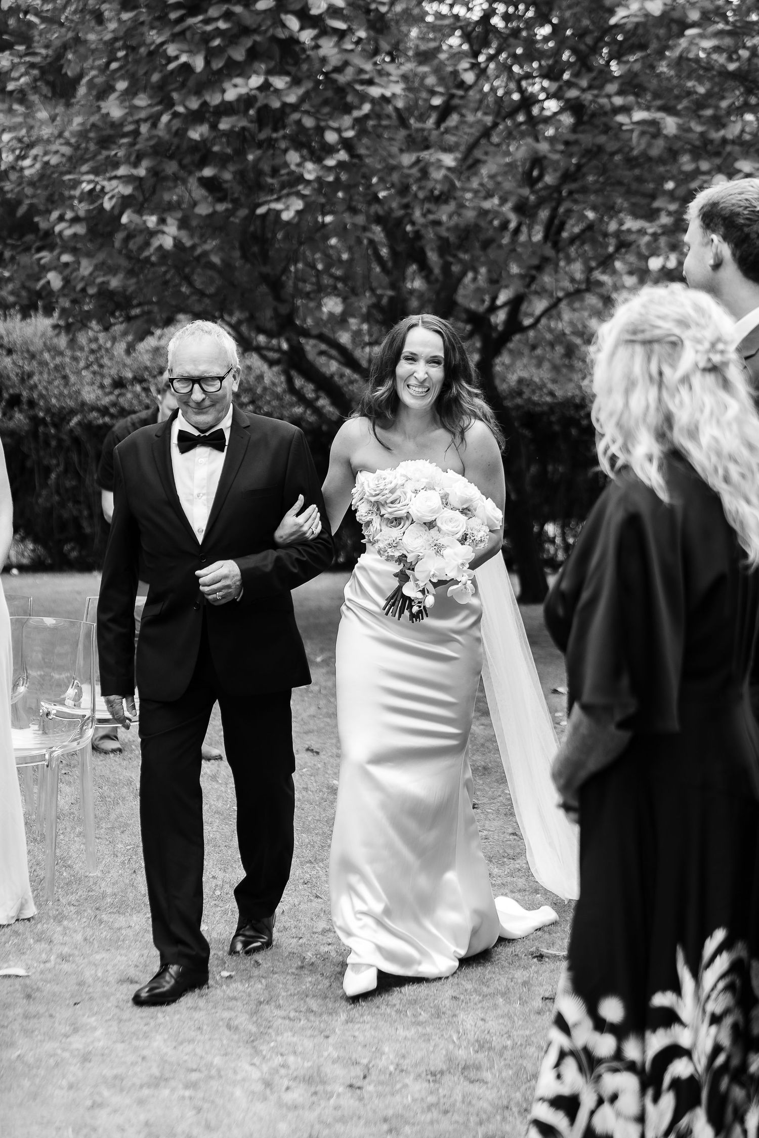 Black and white photo of a smiling bride in a satin gown holding a bouquet, walking down the aisle with an older man in a tuxedo.