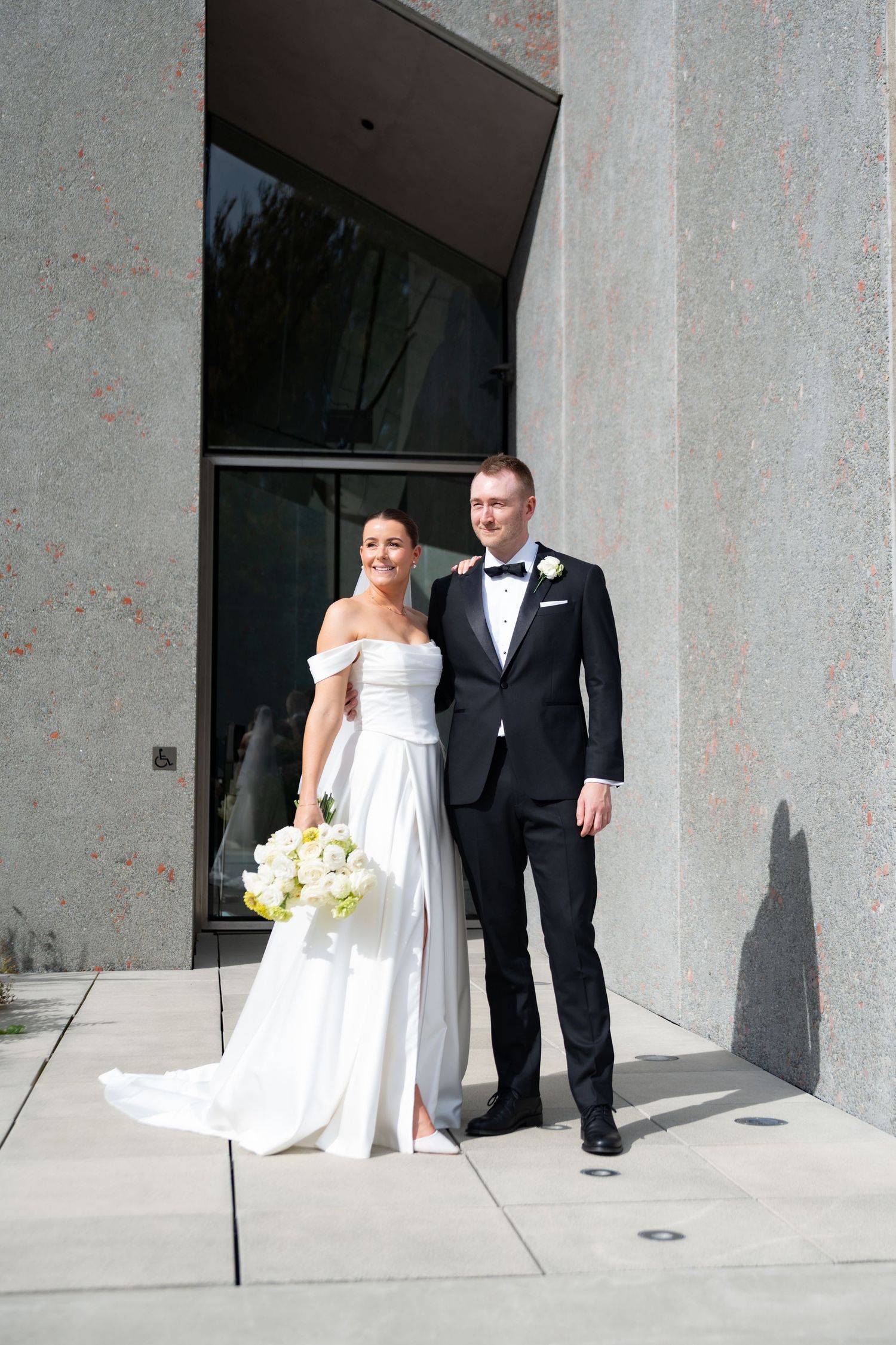 Bride in white off-shoulder gown holding bouquet stands with groom in black tuxedo outside modern stone building.