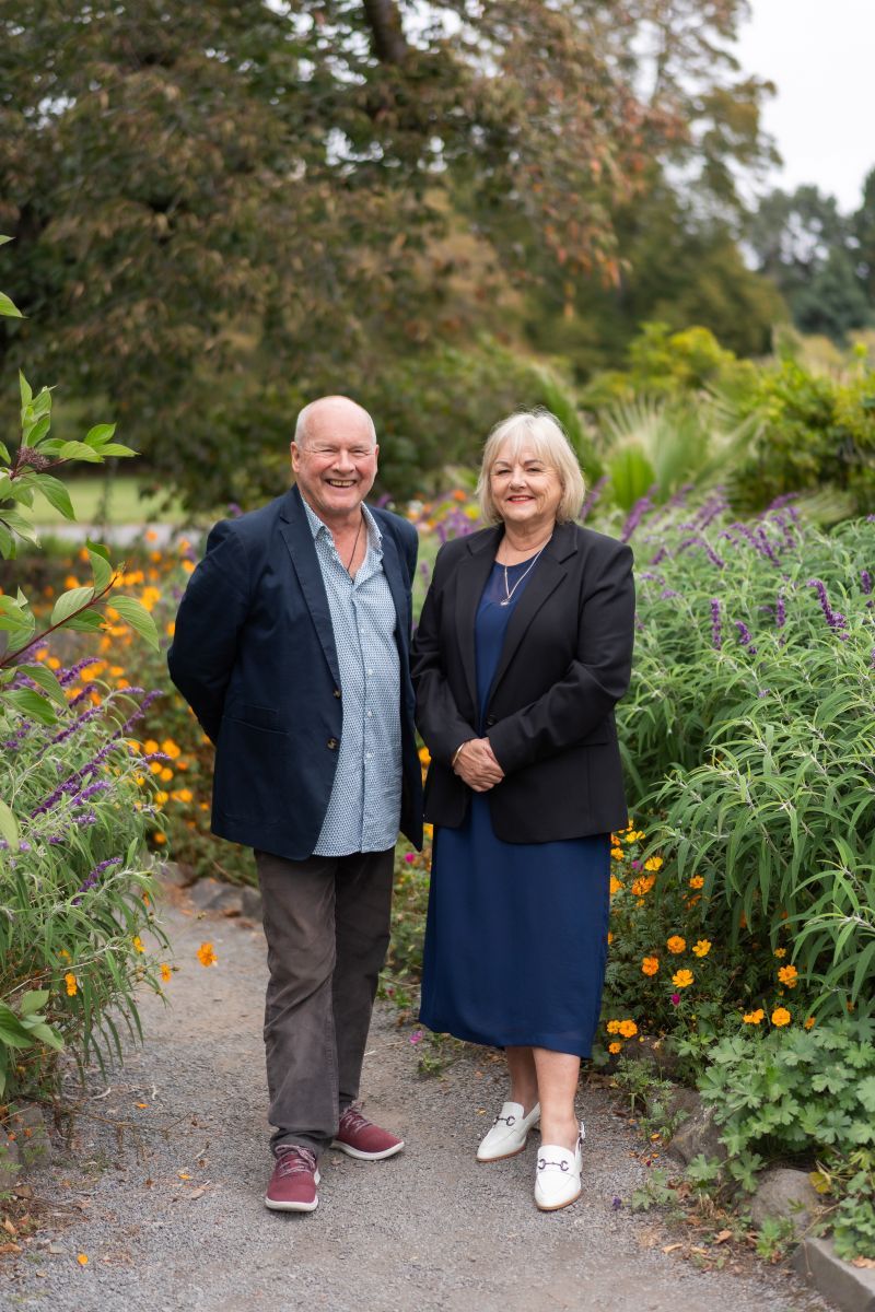 Smiling senior couple standing on a garden path surrounded by colorful flowers and lush greenery.