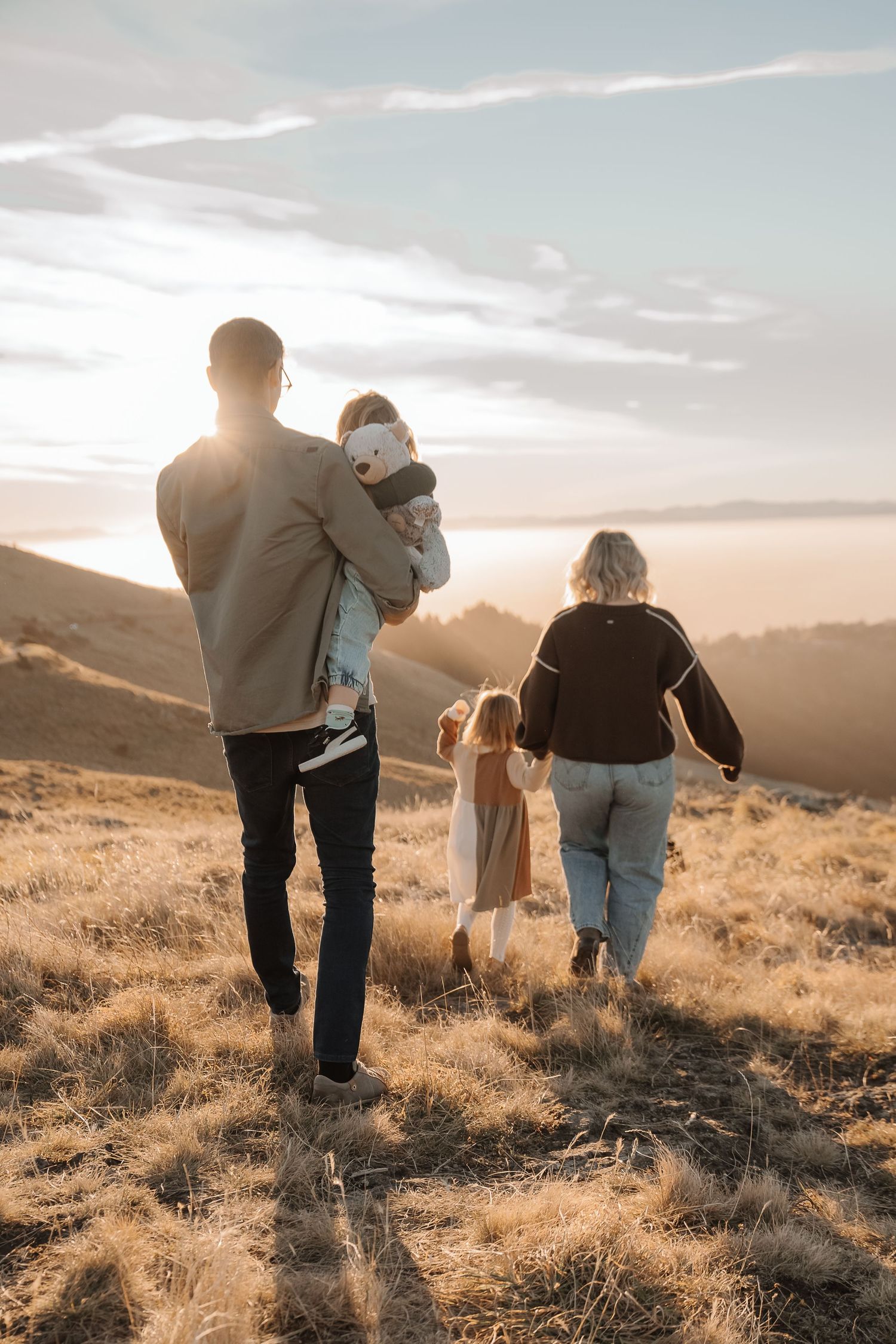 Family of four walking through golden hills at sunset, parents holding children's hands in scenic outdoor landscape.