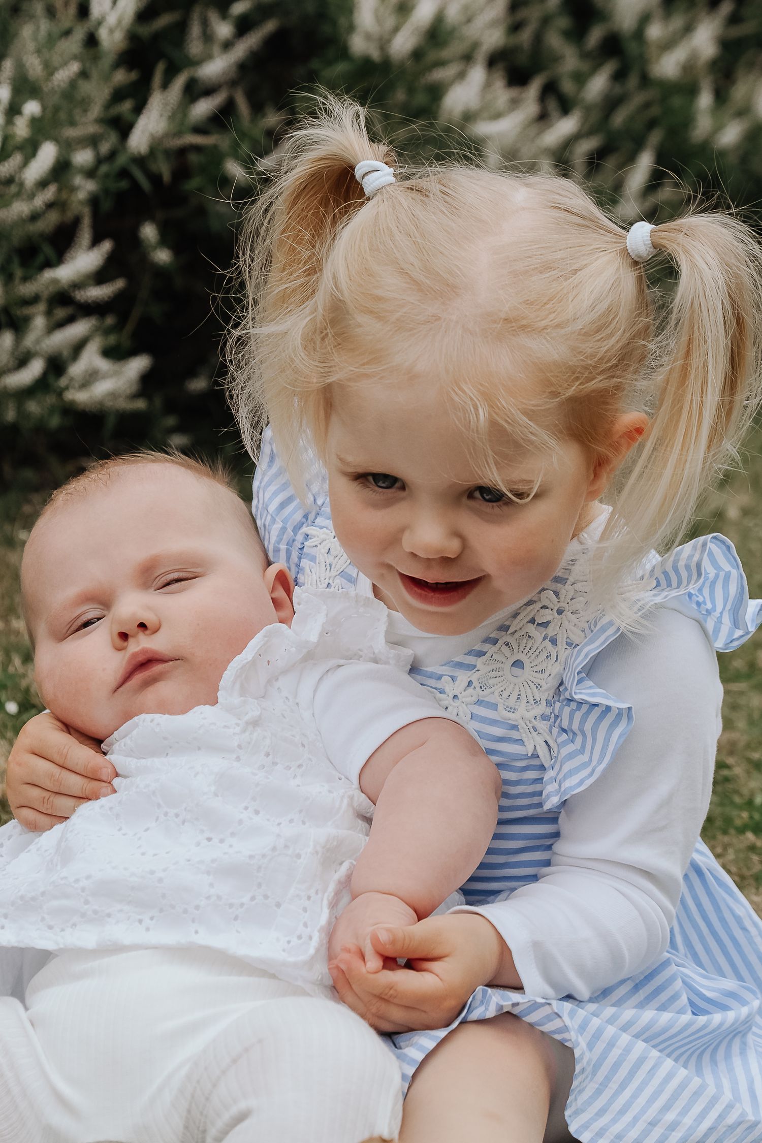 Blonde toddler girl in blue dress holding sleeping newborn baby outdoors in natural setting.