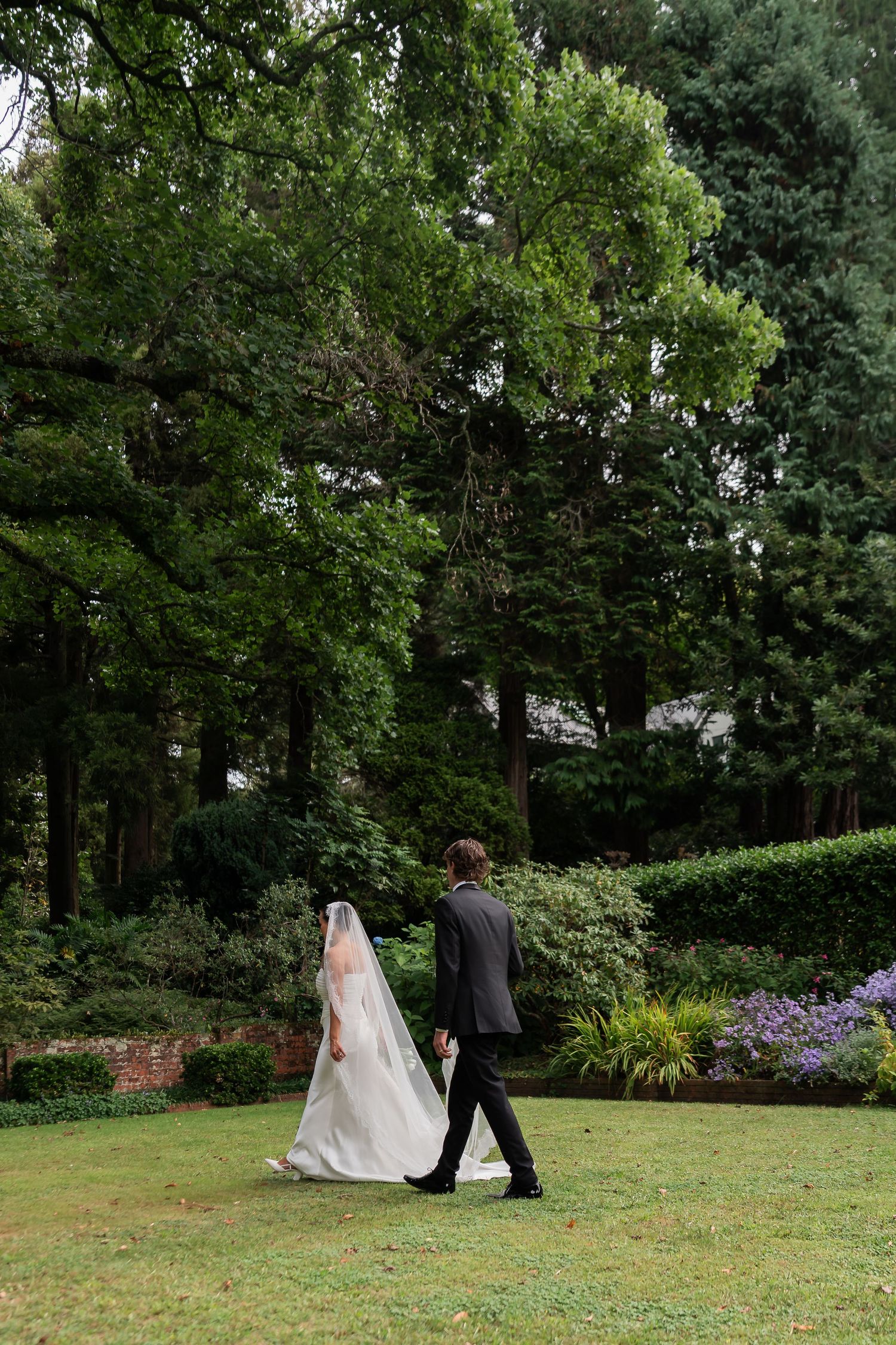 Bride and groom walking hand in hand through a lush green garden, bride wearing a flowing white gown with long veil.