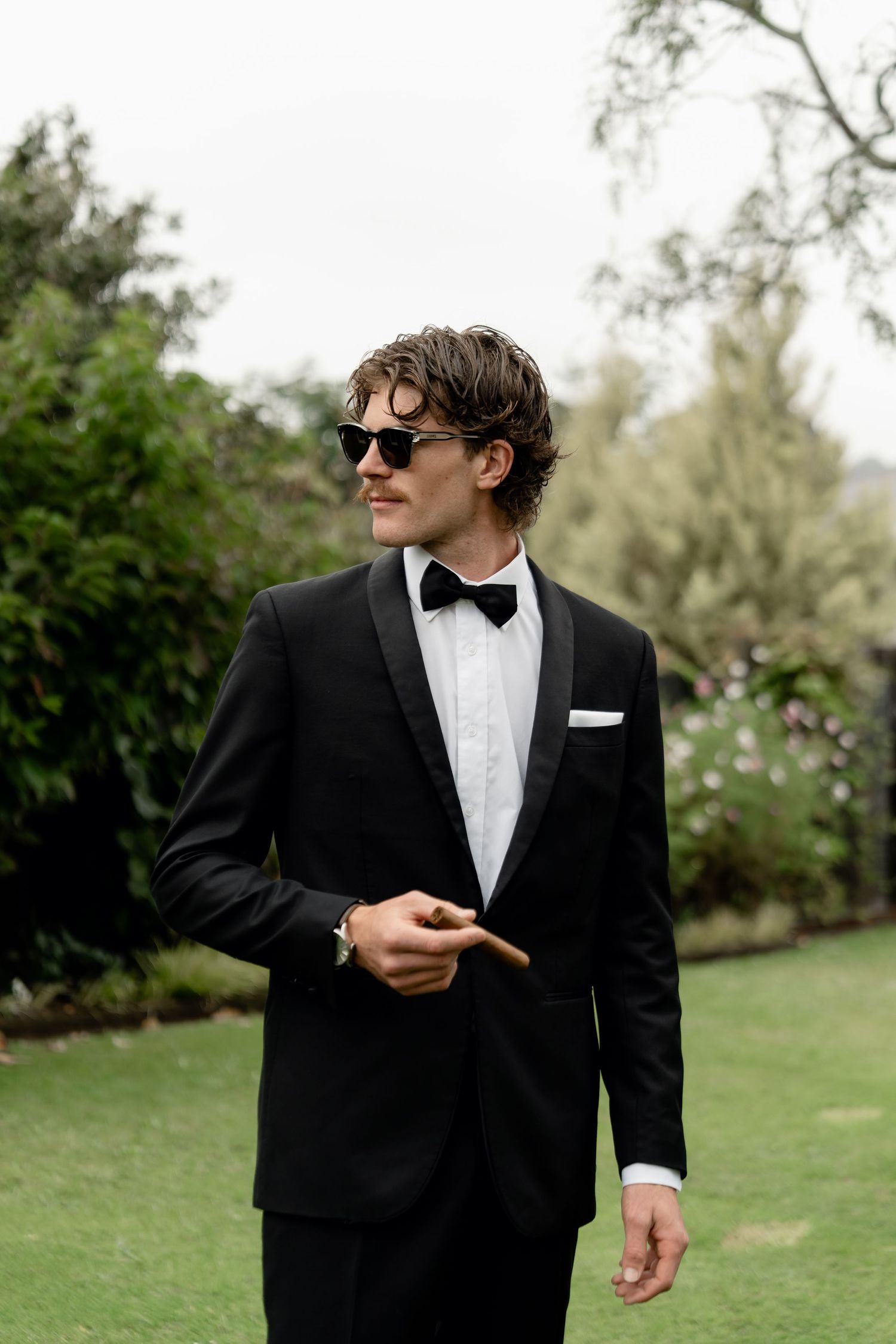 A stylish man in a black tuxedo and bow tie, wearing sunglasses and holding a cigar, standing outdoors.