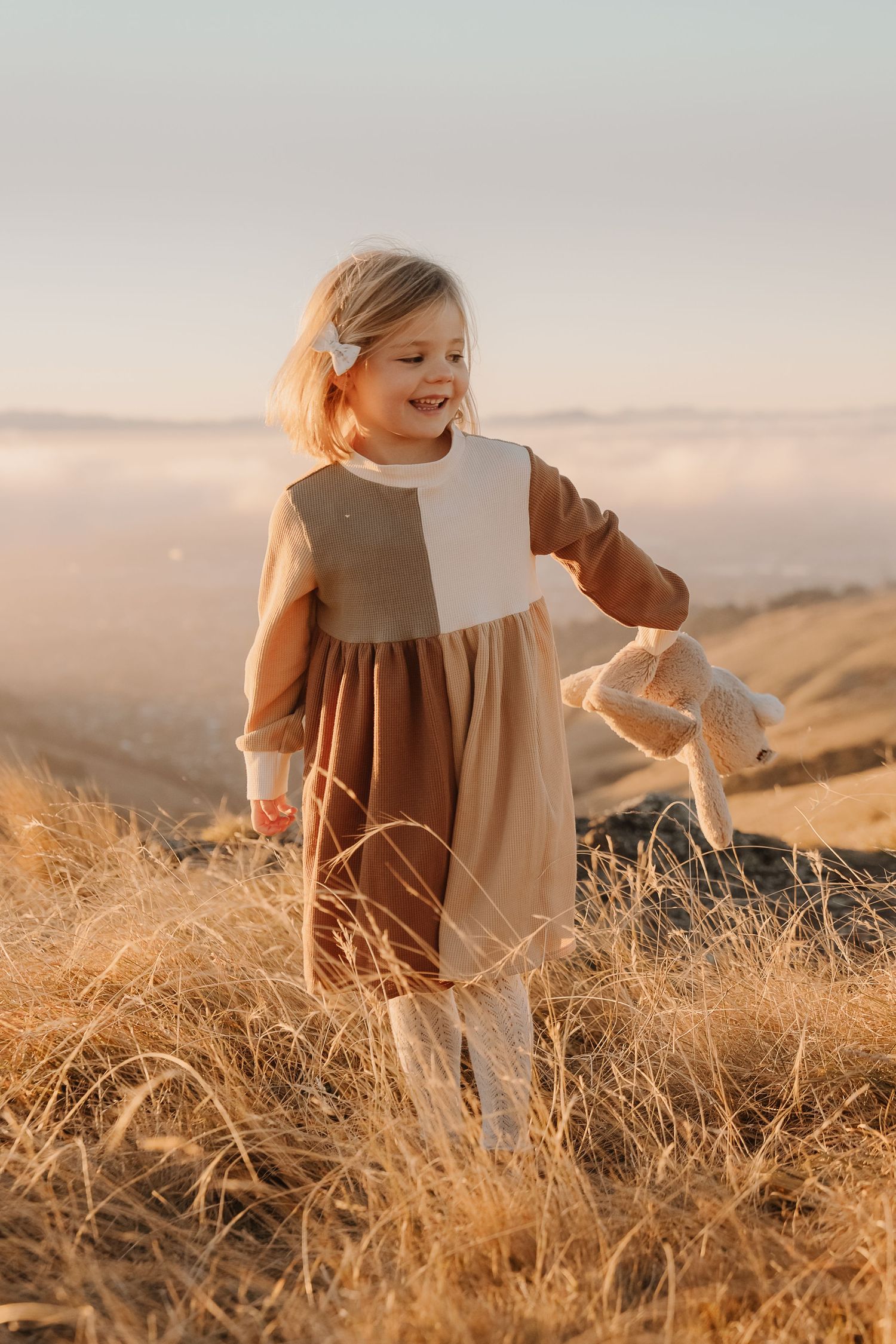Smiling toddler girl in brown colorblock dress walking through golden grass on a hilltop at sunset.