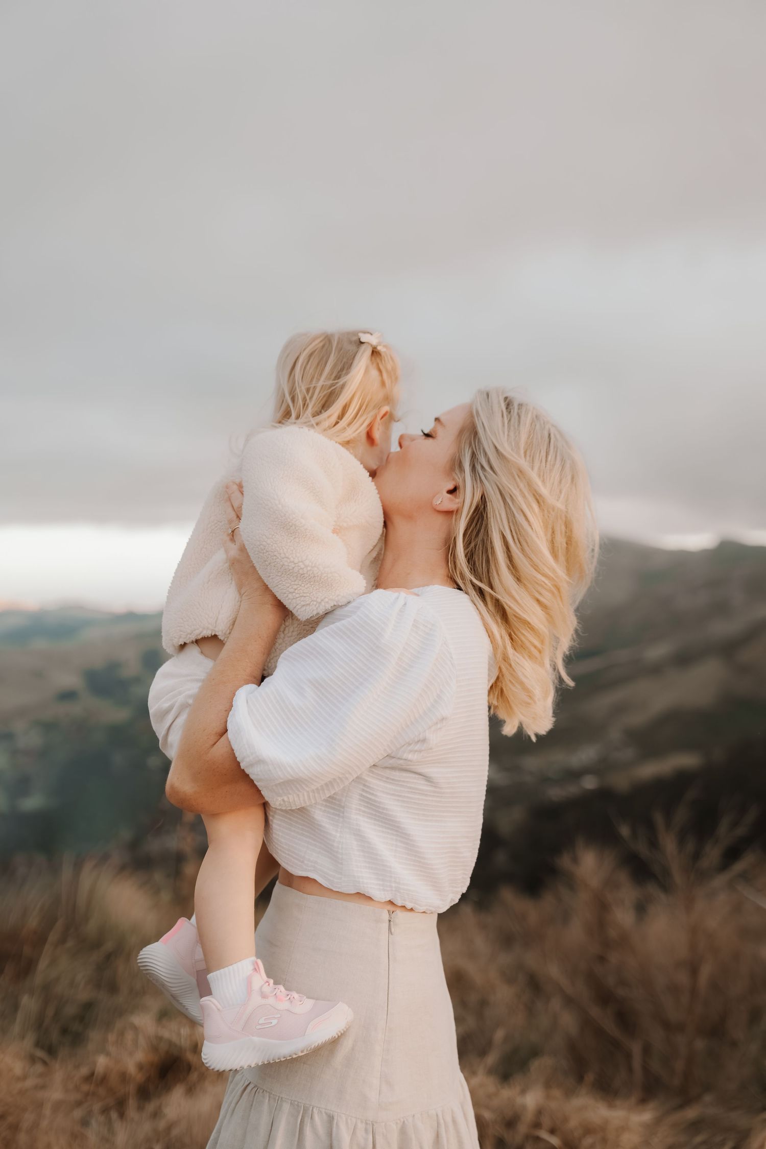 A blonde mother lovingly kisses her toddler daughter outdoors, both wearing soft cream outfits against a scenic mountain backdrop.