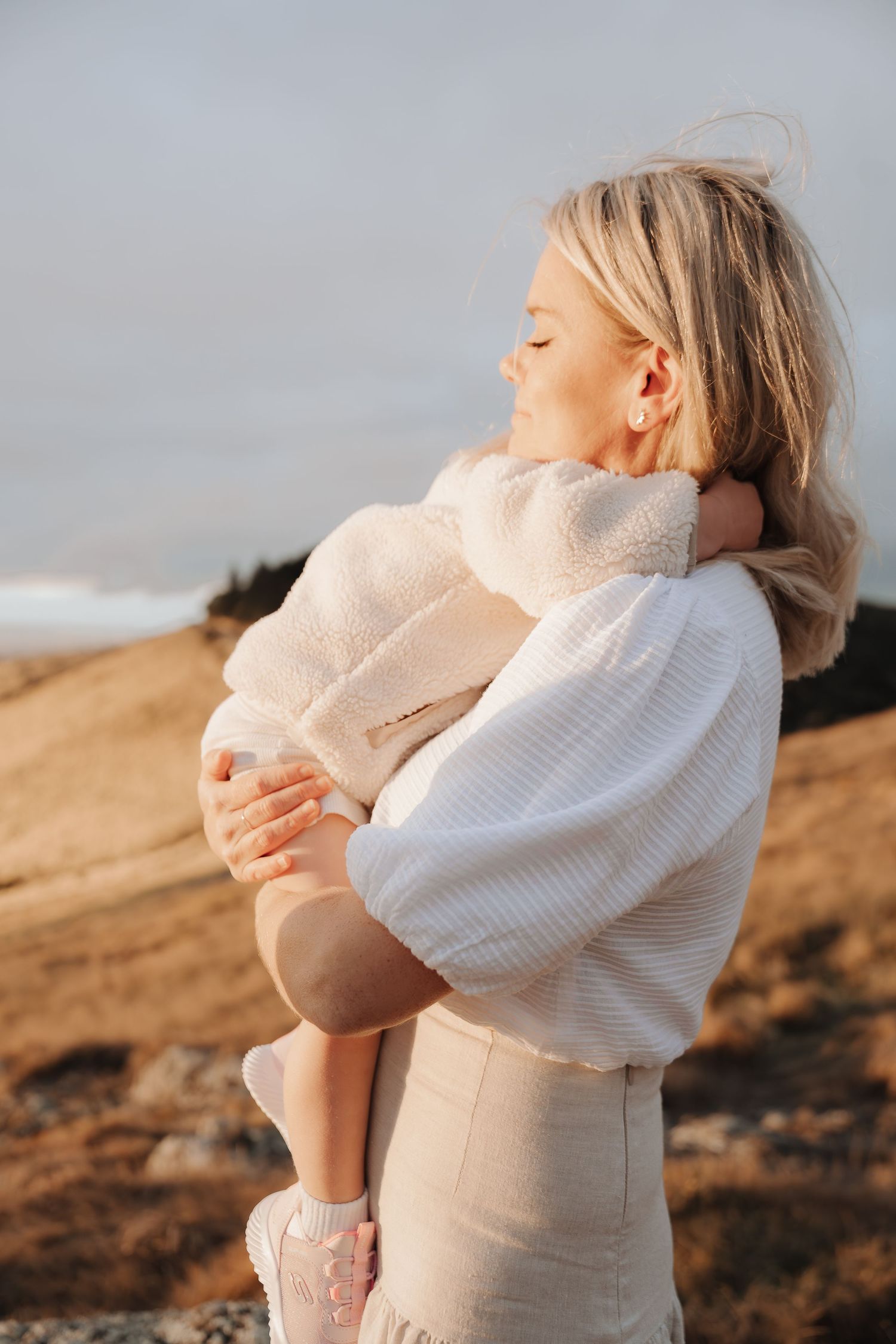 A blonde woman in a white sweater tenderly holds a baby outdoors at golden hour on a scenic hillside.