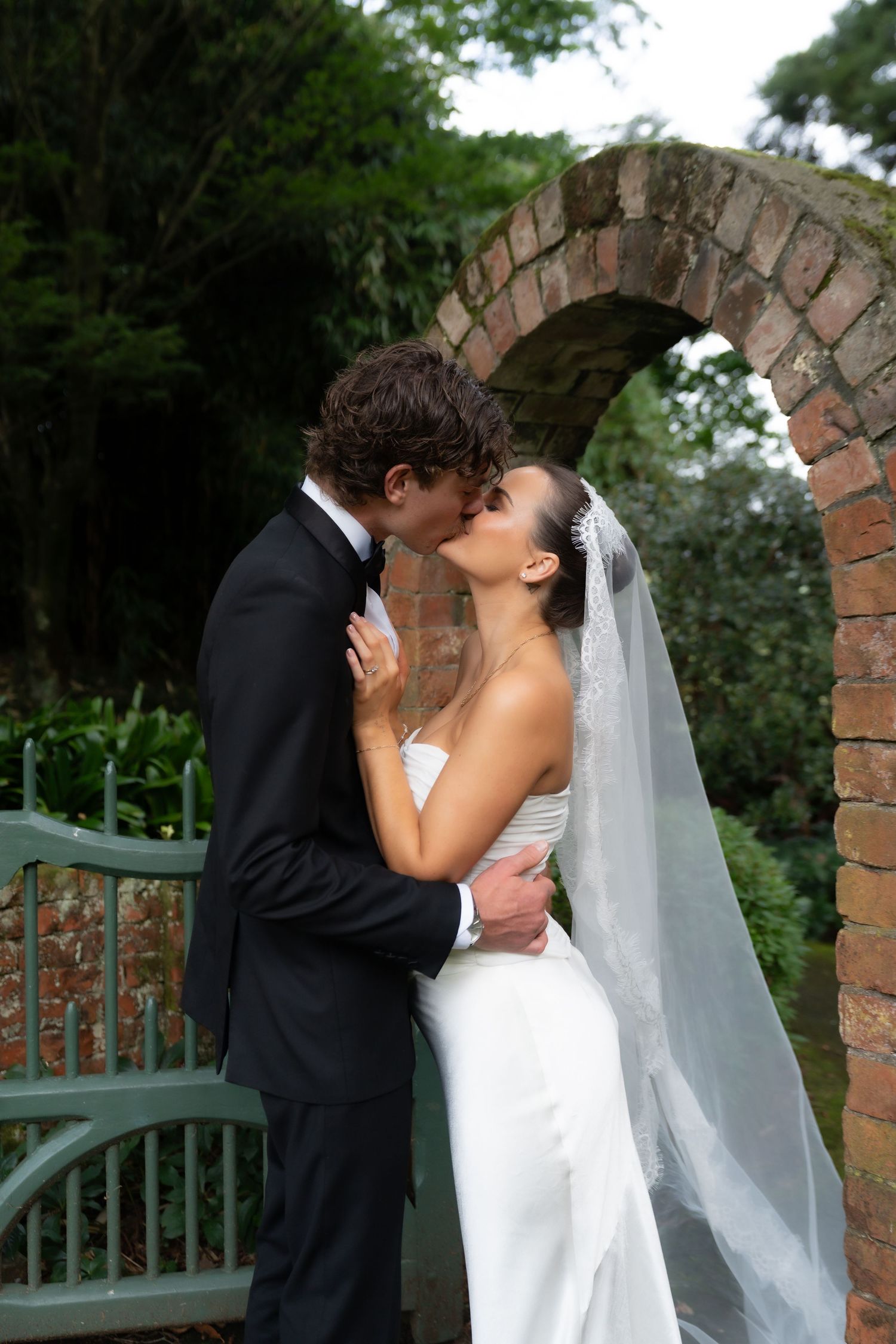 A bride and groom share a romantic kiss under a rustic brick arch surrounded by lush greenery on their wedding day.