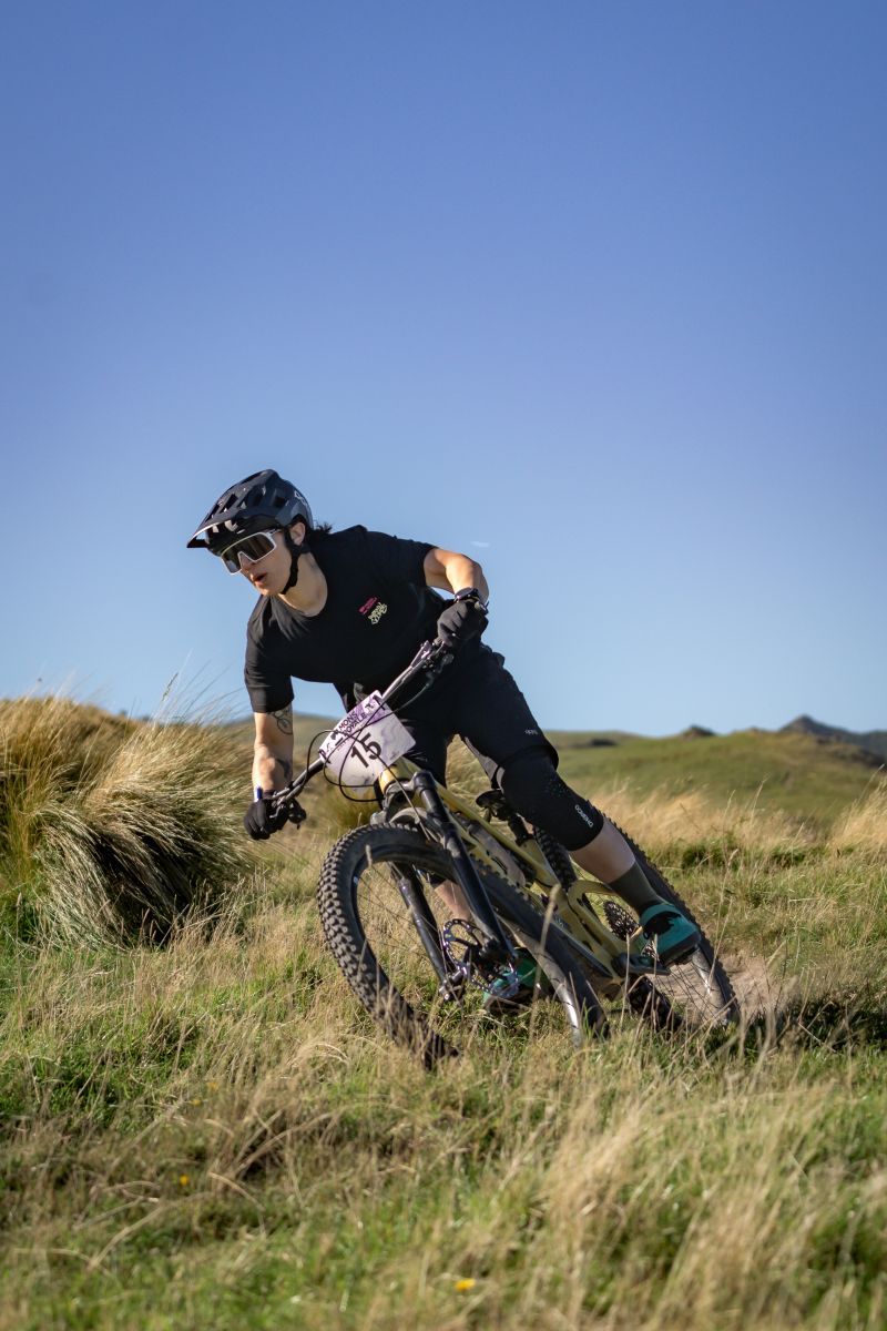 Mountain biker in black gear rides downhill through grassy terrain against blue sky background.