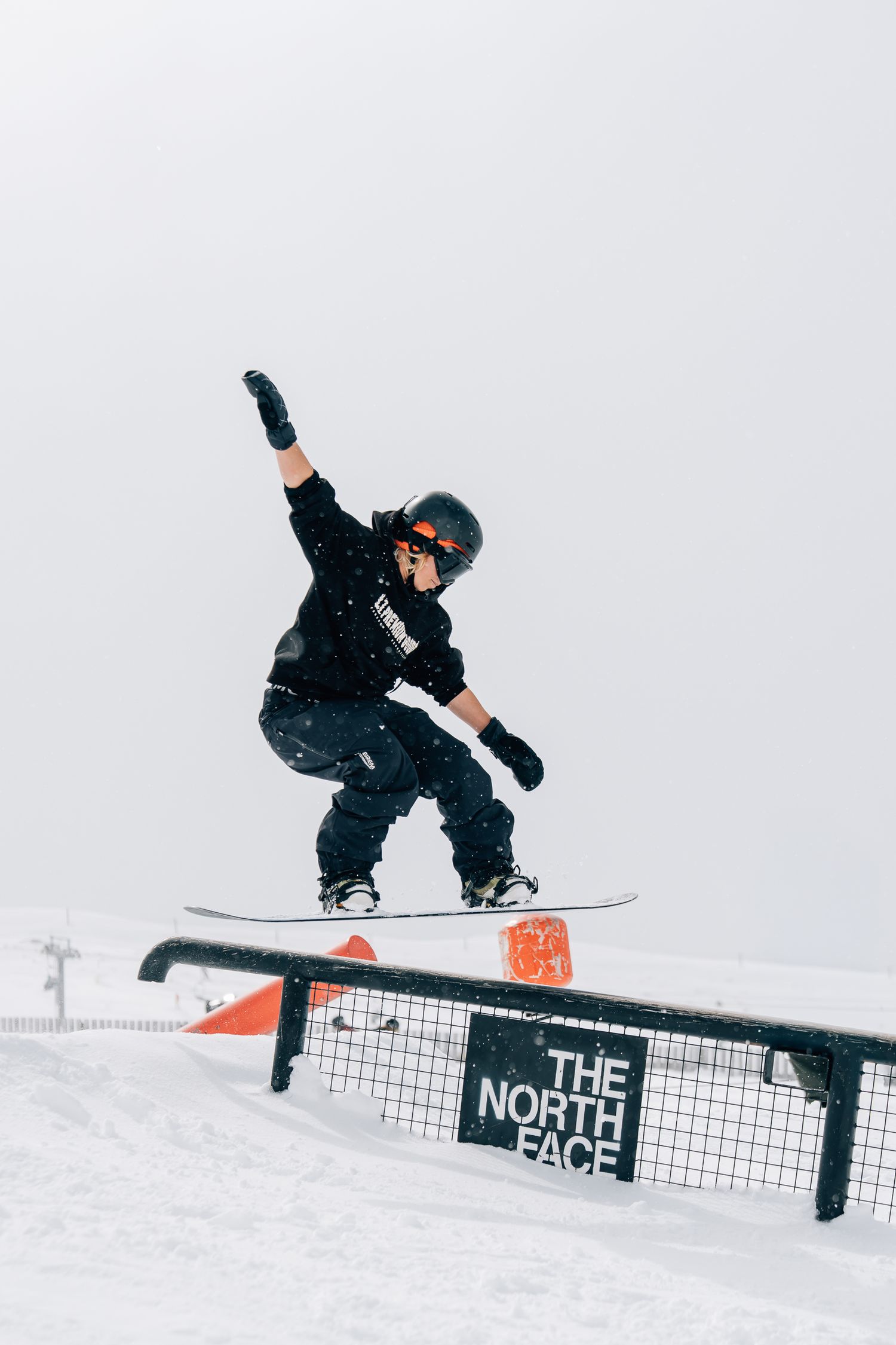 Female snowboarder performing trick sequence on North Face terrain park rail against snowy winter backdrop at Cardrona Alpine Report.