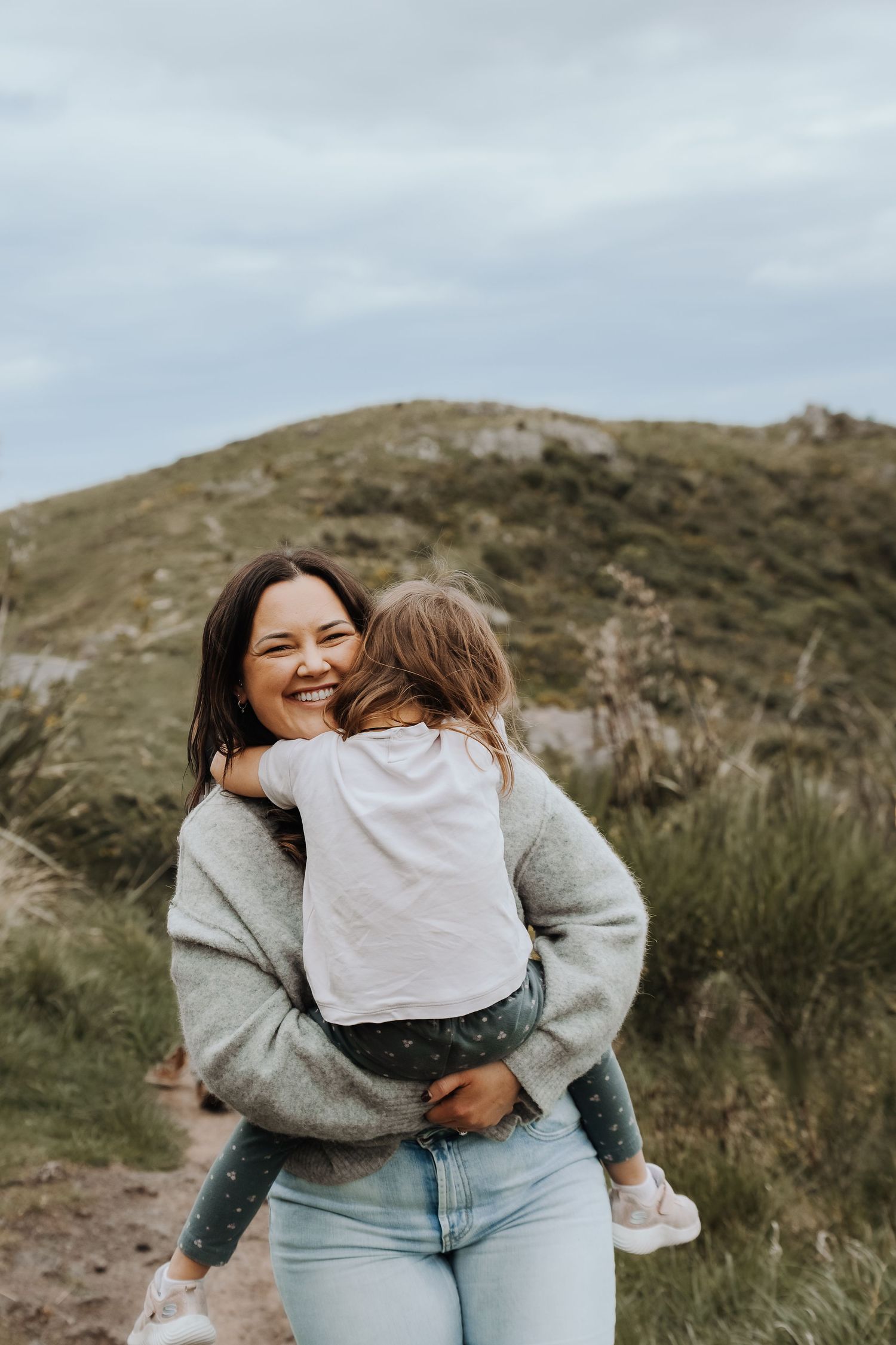 A smiling woman in a gray sweater holds a young child outdoors near green hills under a cloudy sky.