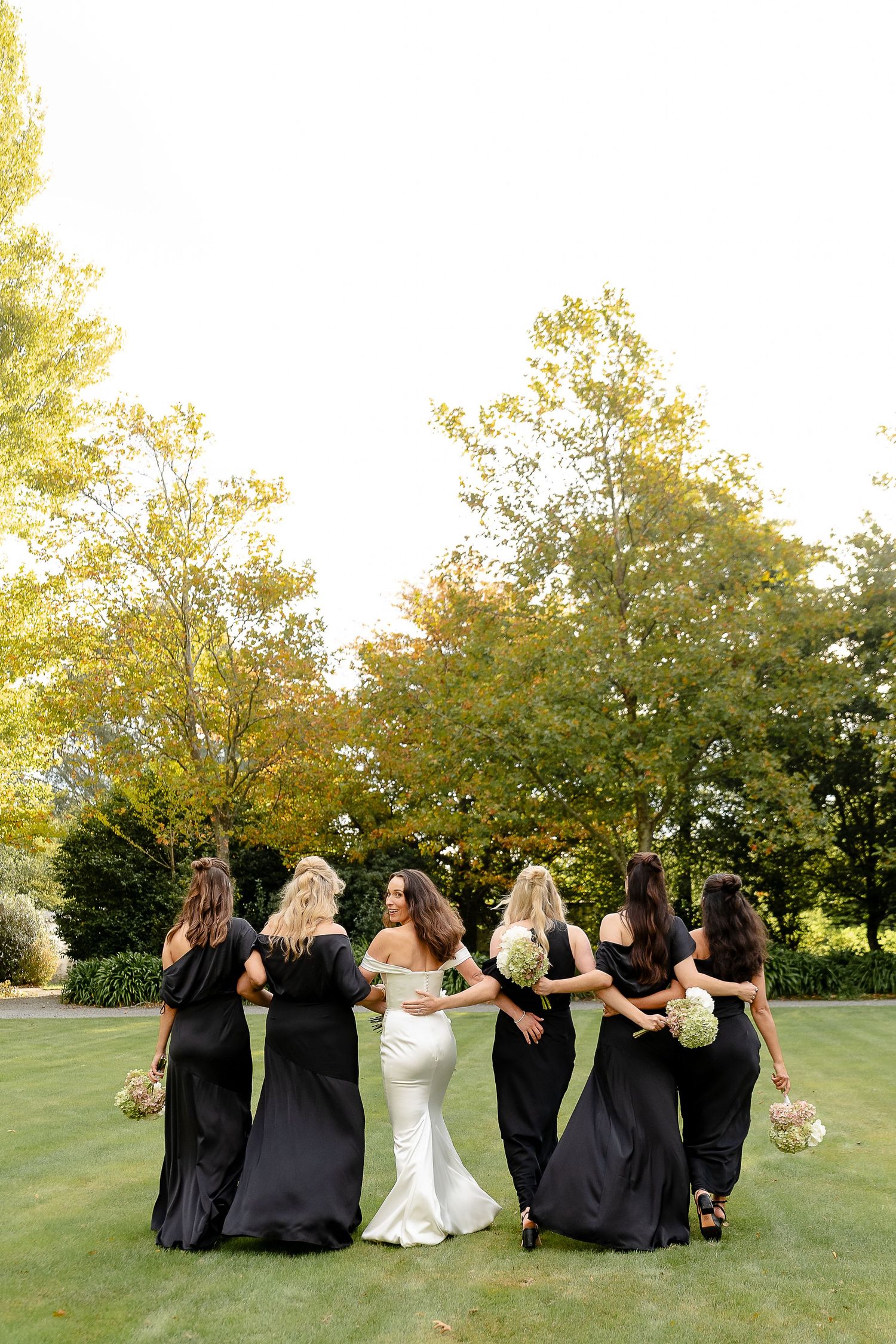 Bride in white gown with bridesmaids in black dresses holding bouquets, posing with backs to camera in lush garden.