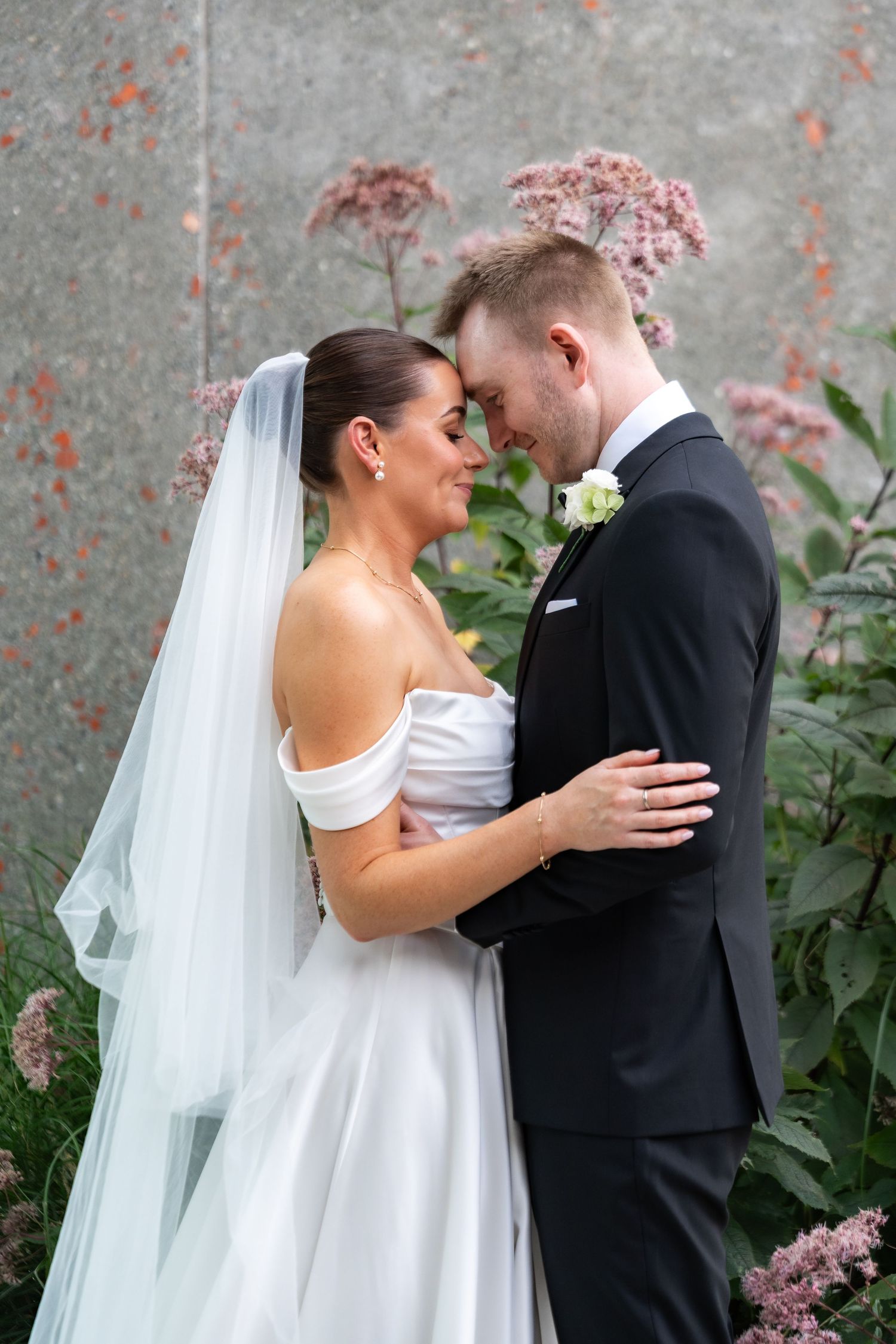 Bride and groom embrace forehead-to-forehead outdoors, wearing white off-shoulder gown with veil and black tuxedo.