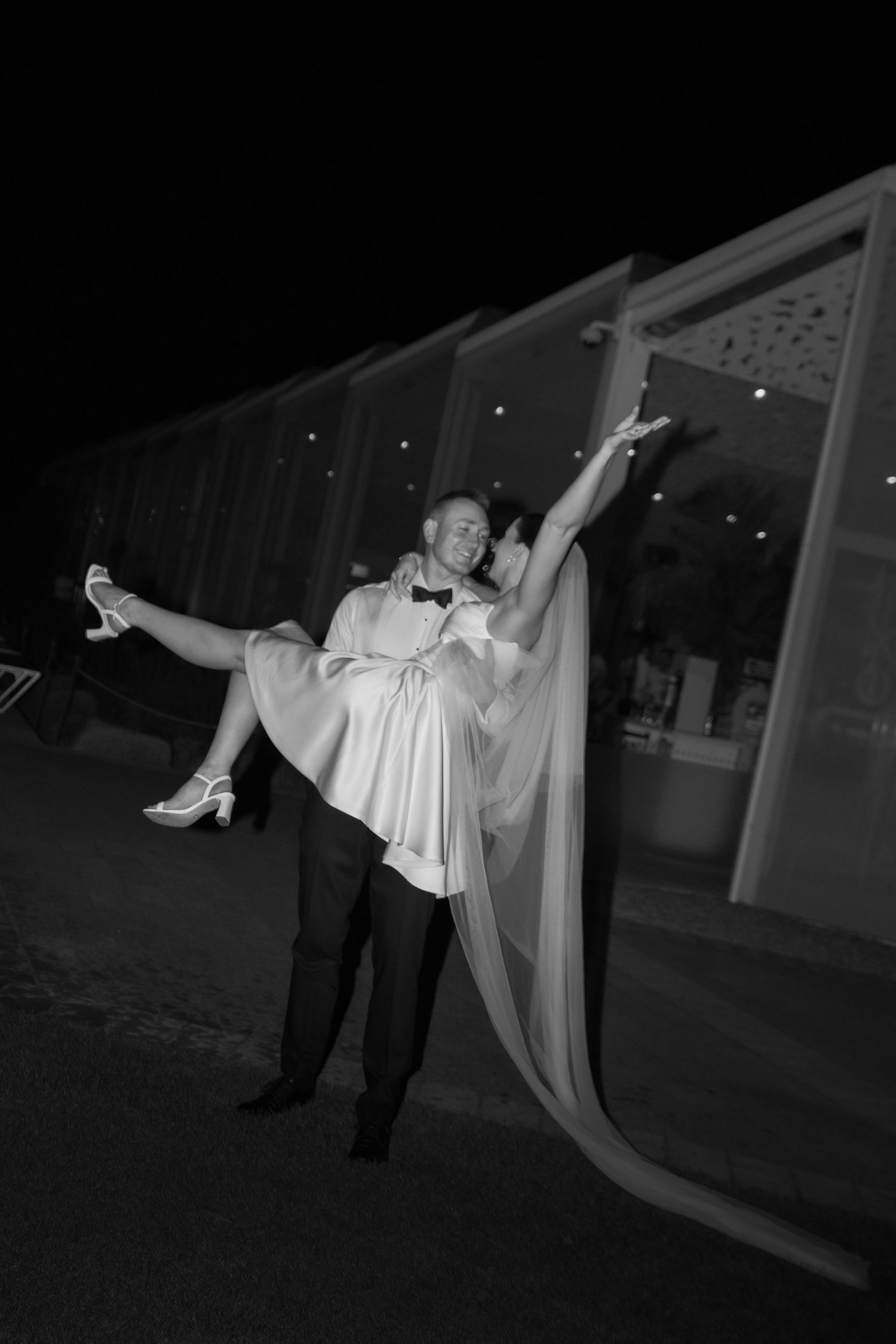 Black and white wedding photo of groom lifting bride in white dress outdoors at night.