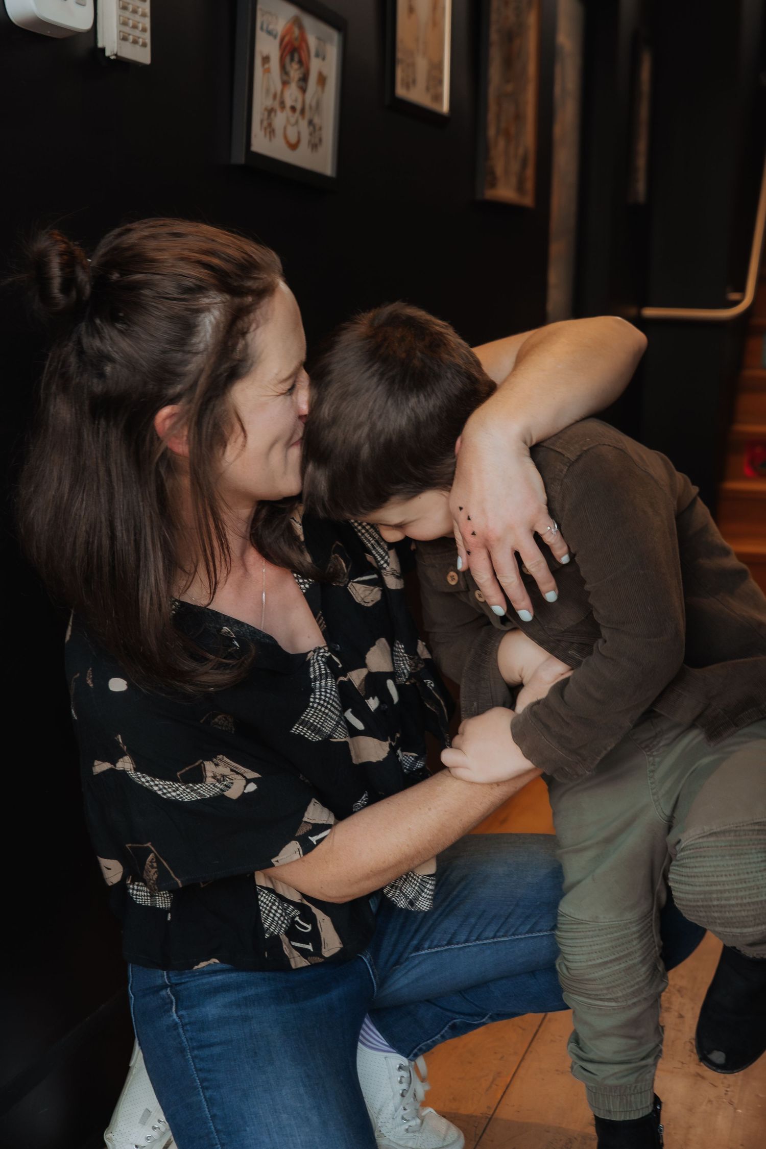 A woman with dark hair smiles warmly while hugging a young boy in a tattoo shop.