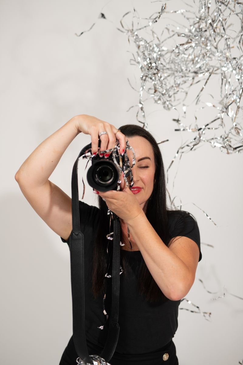 Woman in black outfit photographing with DSLR camera, silver tinsel decoration in background.