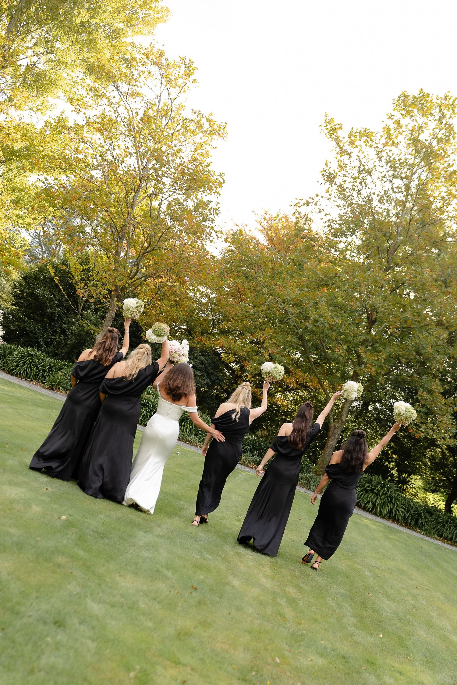 Bride in white gown with bridesmaids in black dresses raising bouquets on green lawn surrounded by autumn trees.
