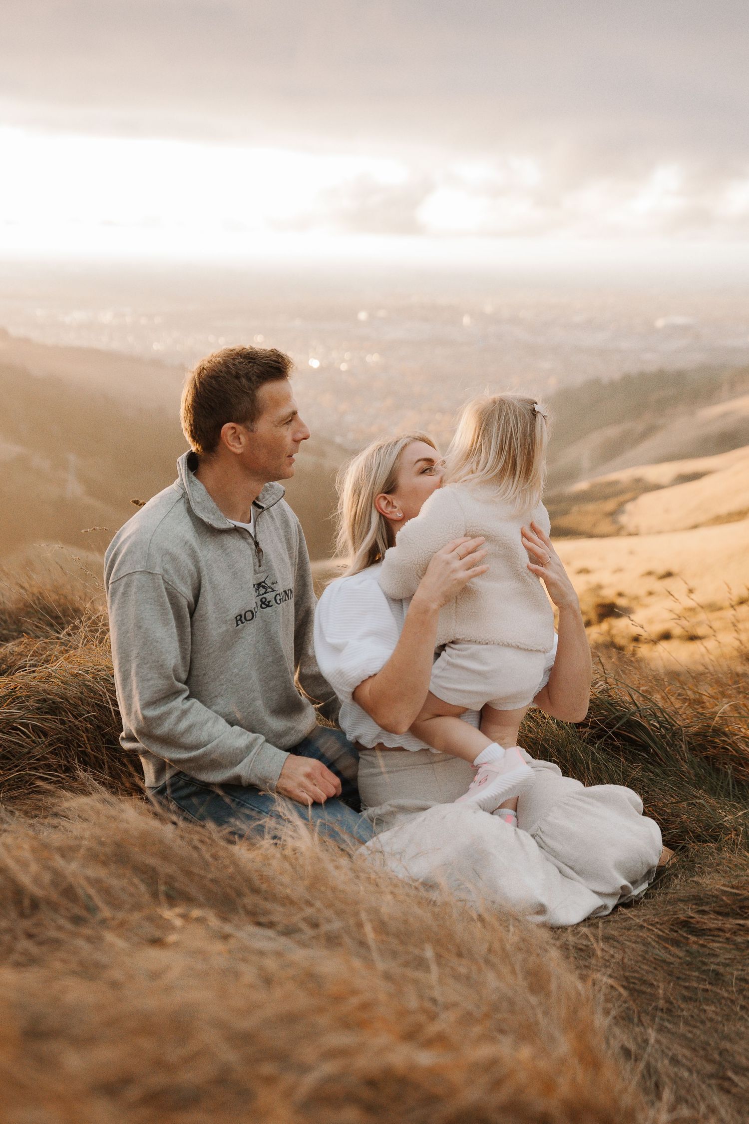 Family sitting on a hillside at sunset, parents lovingly holding their young child with a scenic valley view.