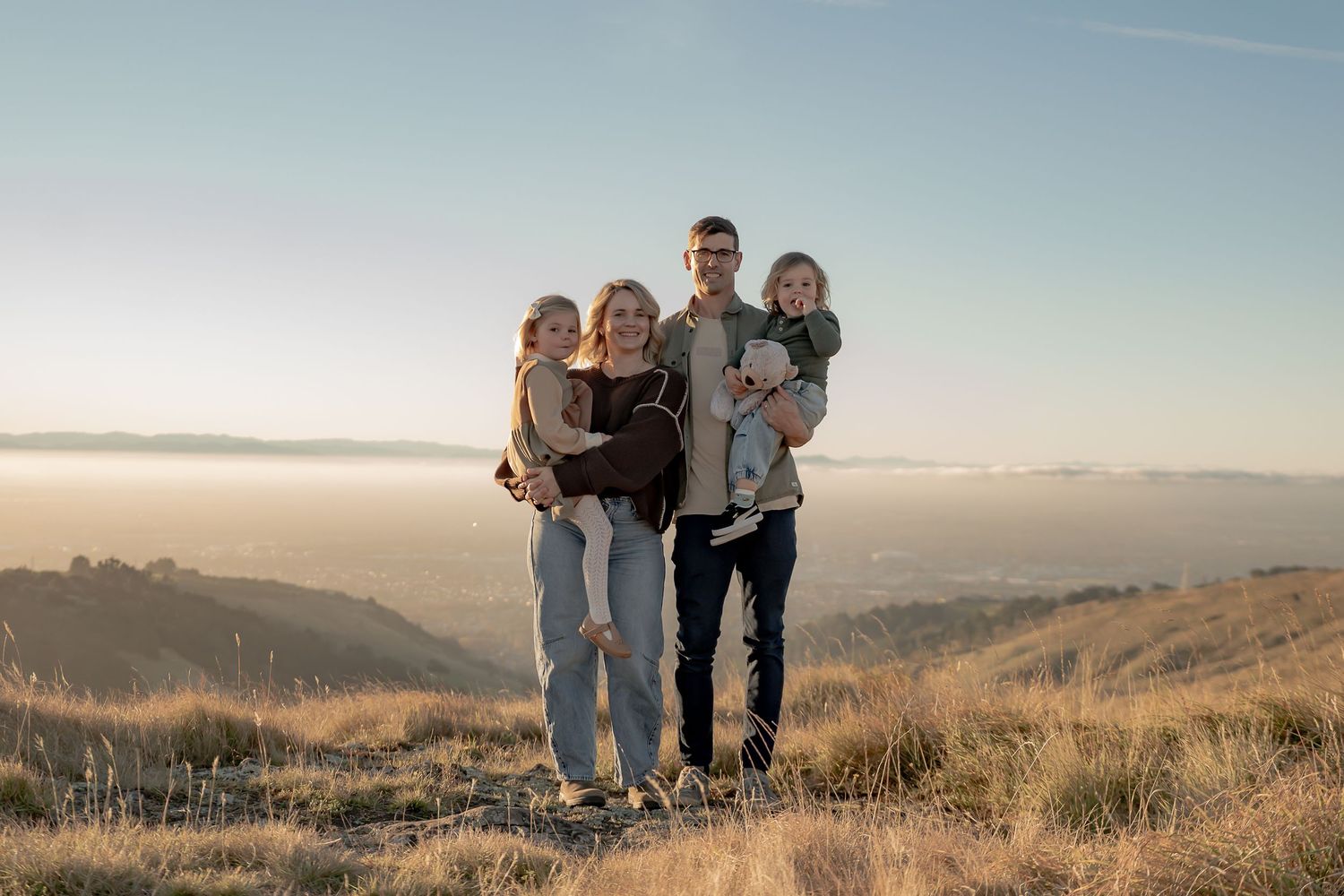 Family of four posing on a hilltop at golden hour with scenic valley and misty mountains in the background.