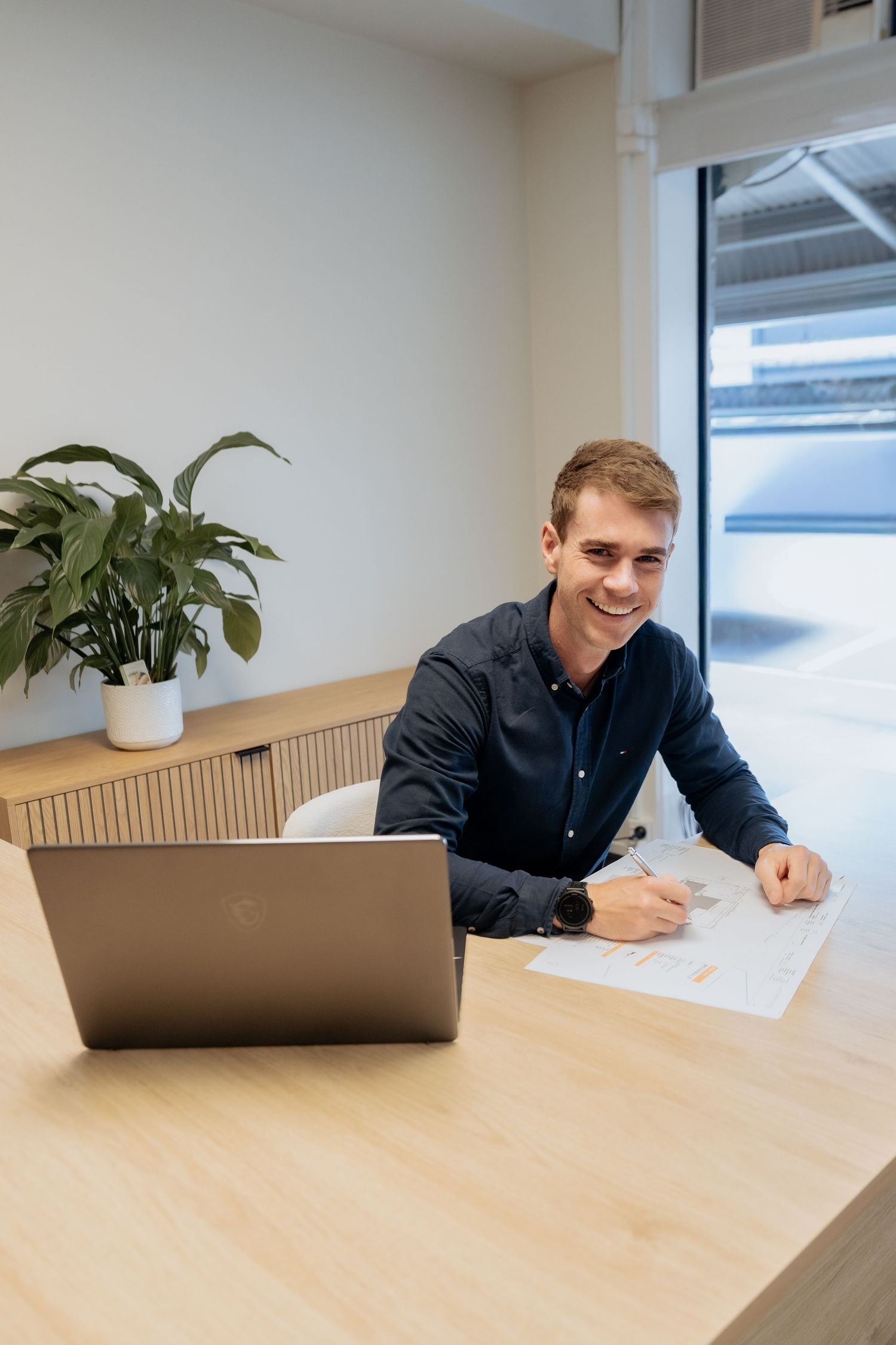 Smiling young businessman in dark shirt working at wooden desk with laptop and documents in modern office.