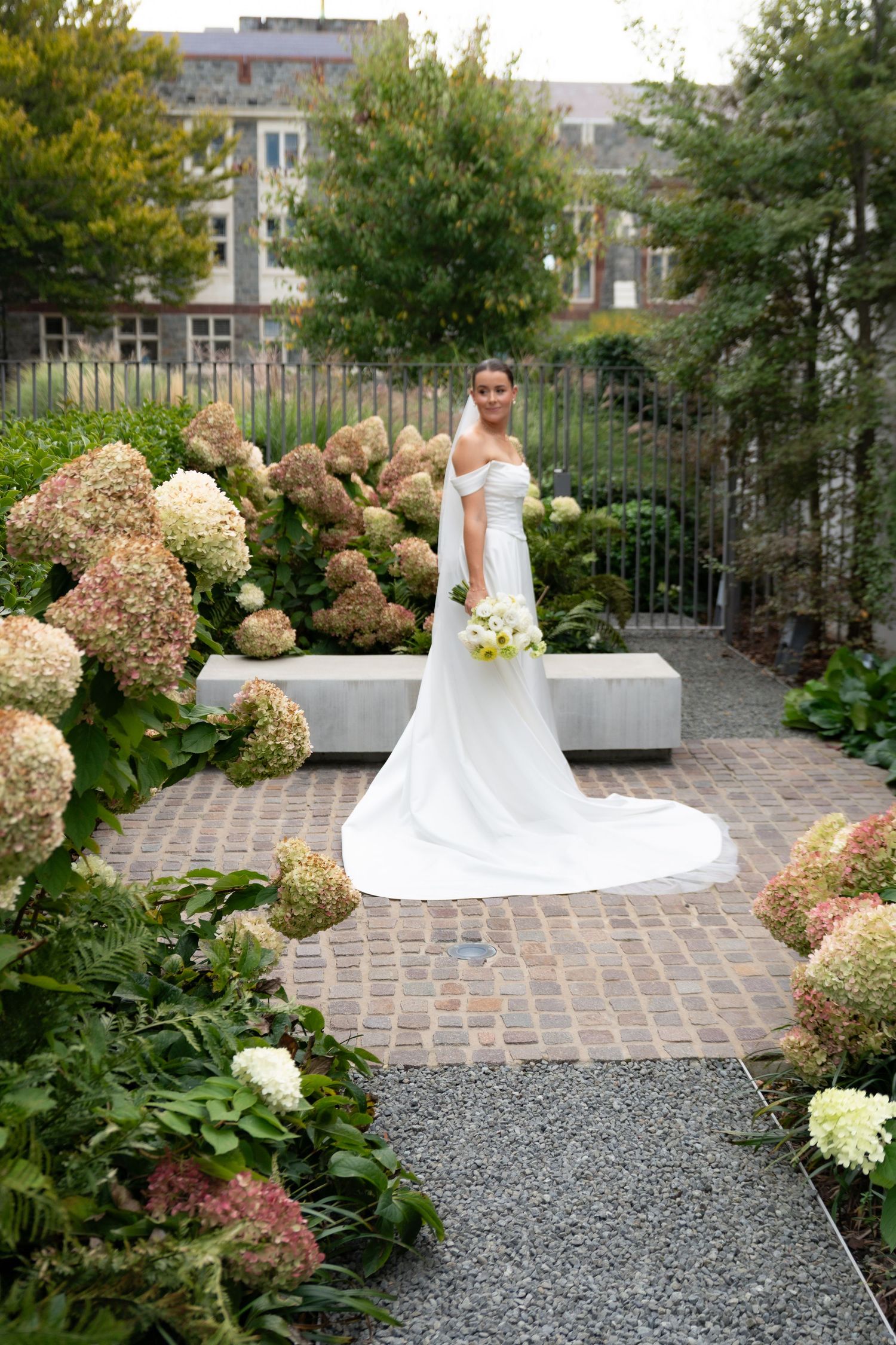Bride in white off-shoulder gown with train stands among blooming hydrangeas on brick path in lush garden.