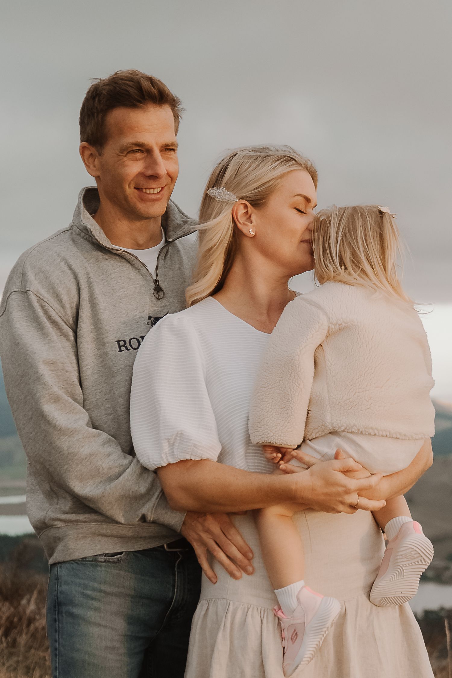 A smiling family with a man in a gray sweatshirt, a blonde woman kissing their young child outdoors.