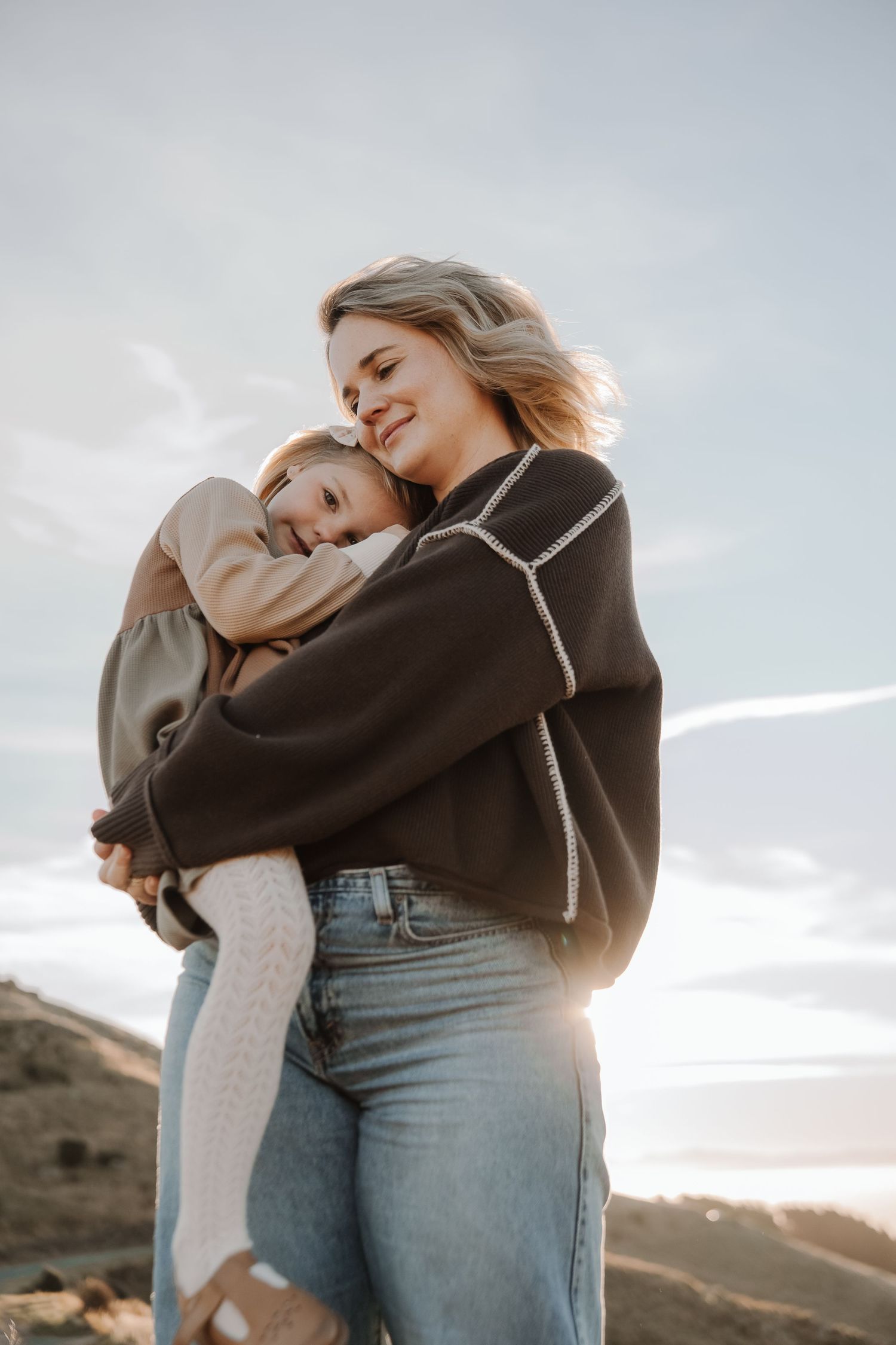 A smiling woman in a dark sweater and jeans lovingly hugs a young girl outdoors under a bright sky.