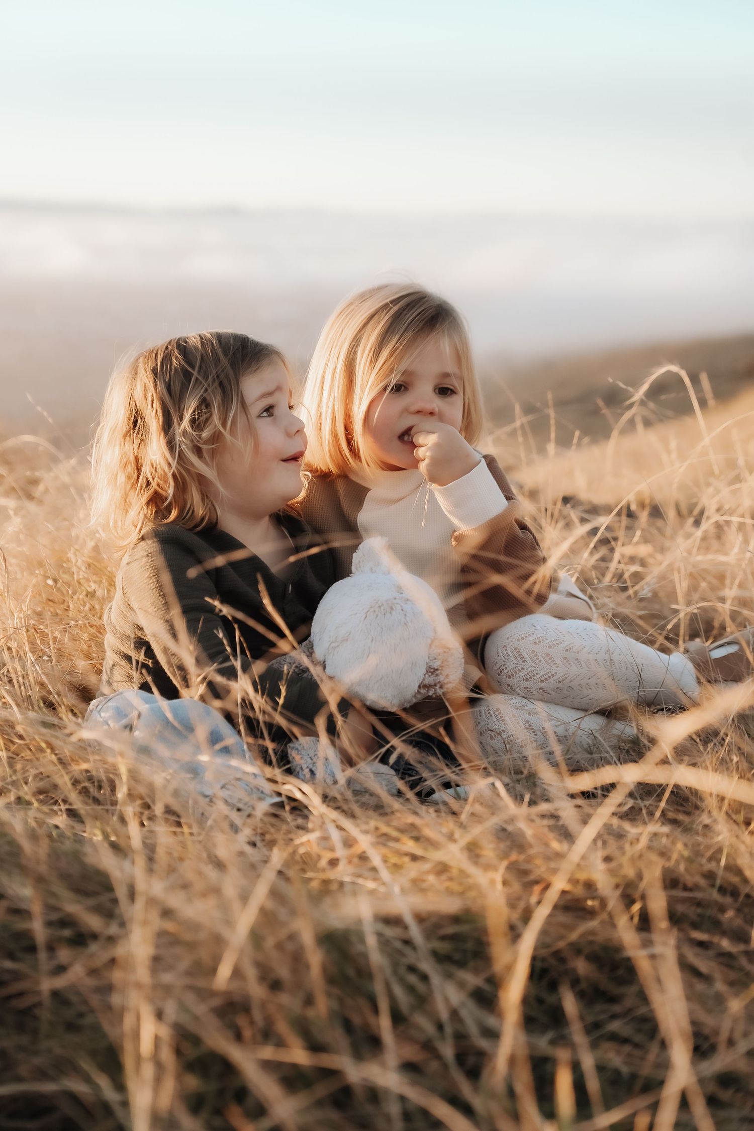 Two young children sitting in golden dry grass field, wrapped in cozy blankets during a warm sunset outdoors.