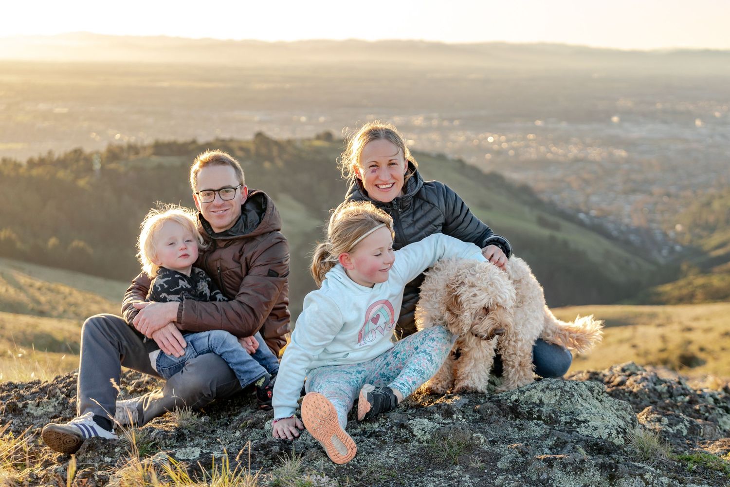 Family and dog laying on grassy hilltop during golden sunset overlooking scenic mountain landscape.