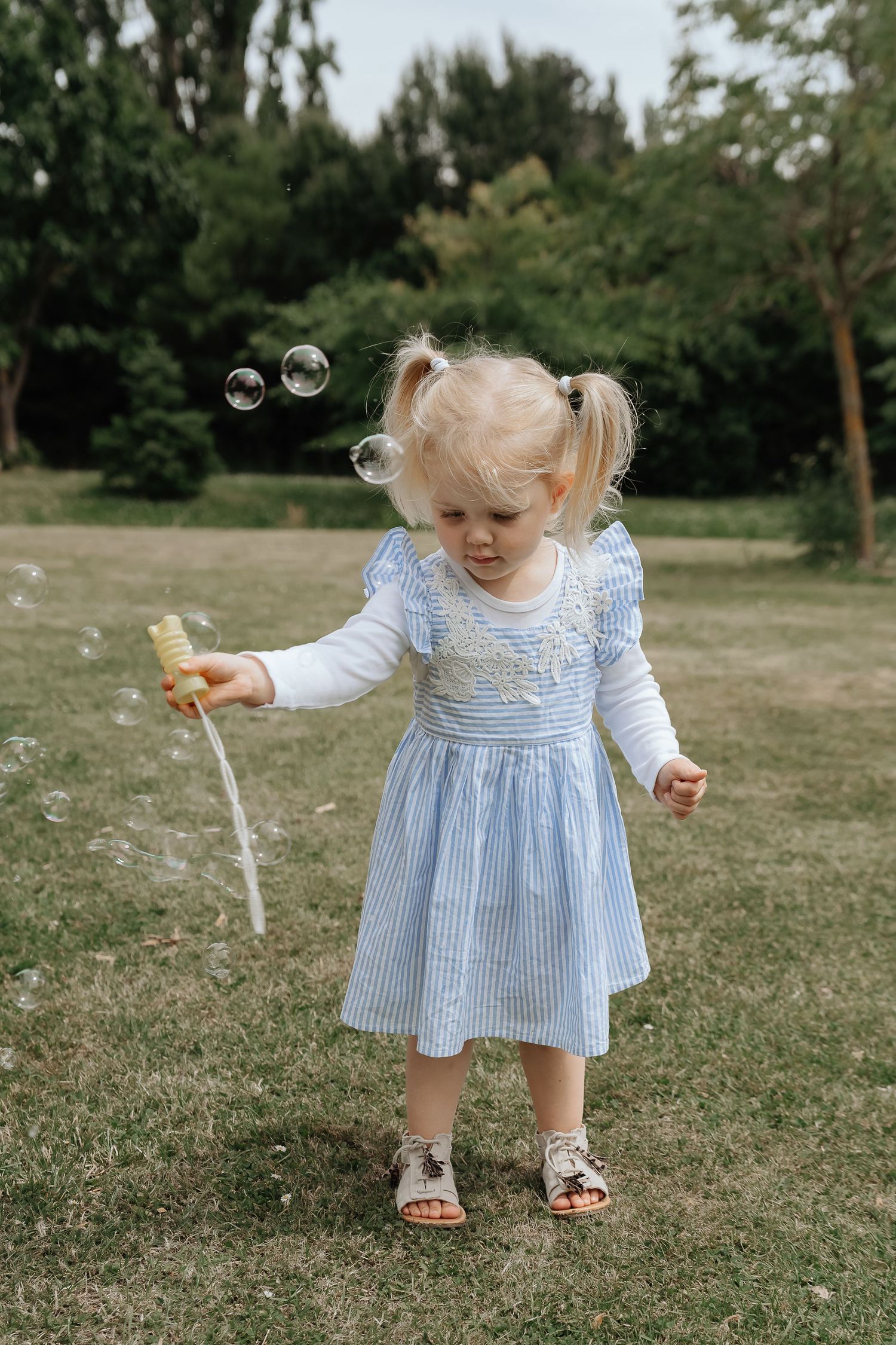 Toddler girl in blue striped dress blowing bubbles in a green park, holding a bubble wand.