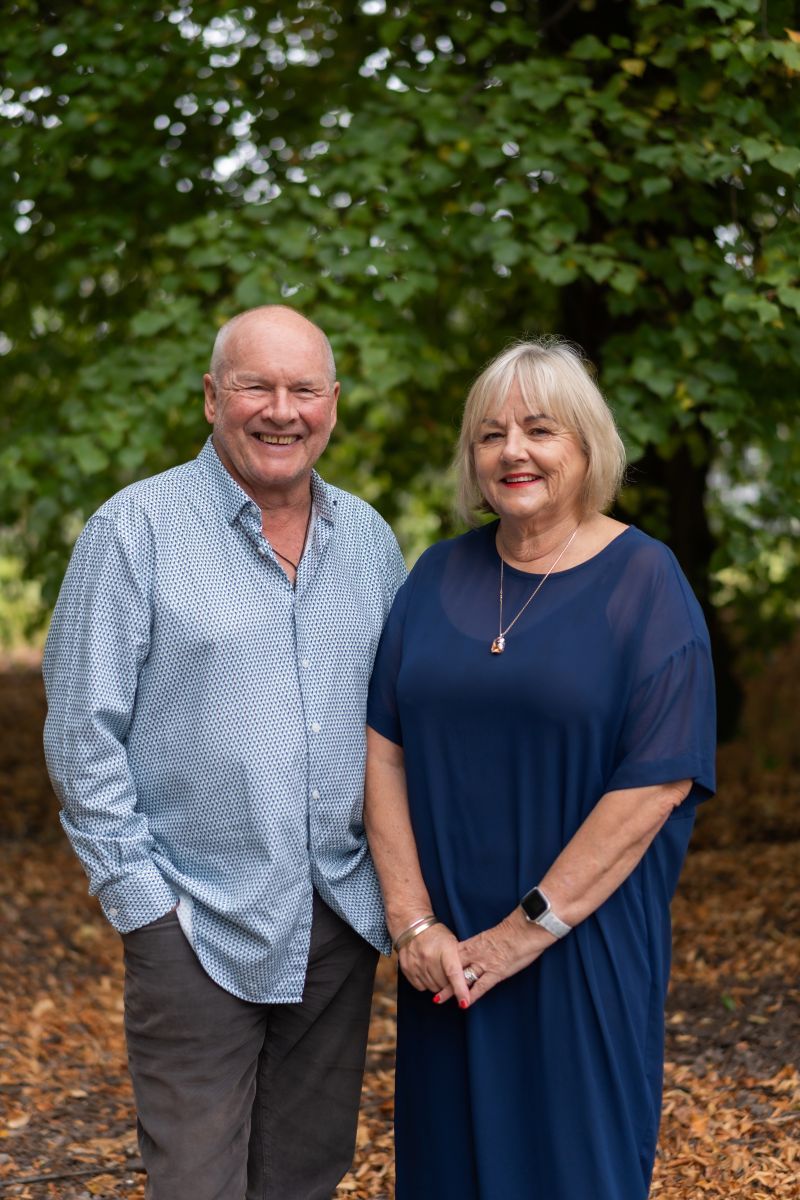 Couple in blue and light colored clothing pose together outdoors among autumn leaves and green foliage.