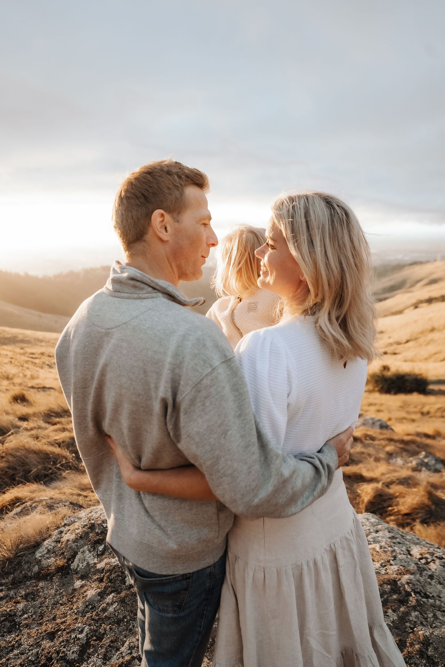 A couple embraces with their child at golden hour in rolling hills, wearing neutral tones in a warm family portrait.